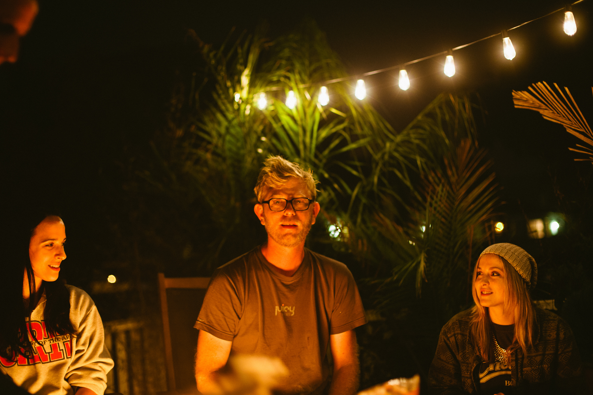 Man with glasses chatting with two women around an outdoor fire pit under glowing string lights at night.