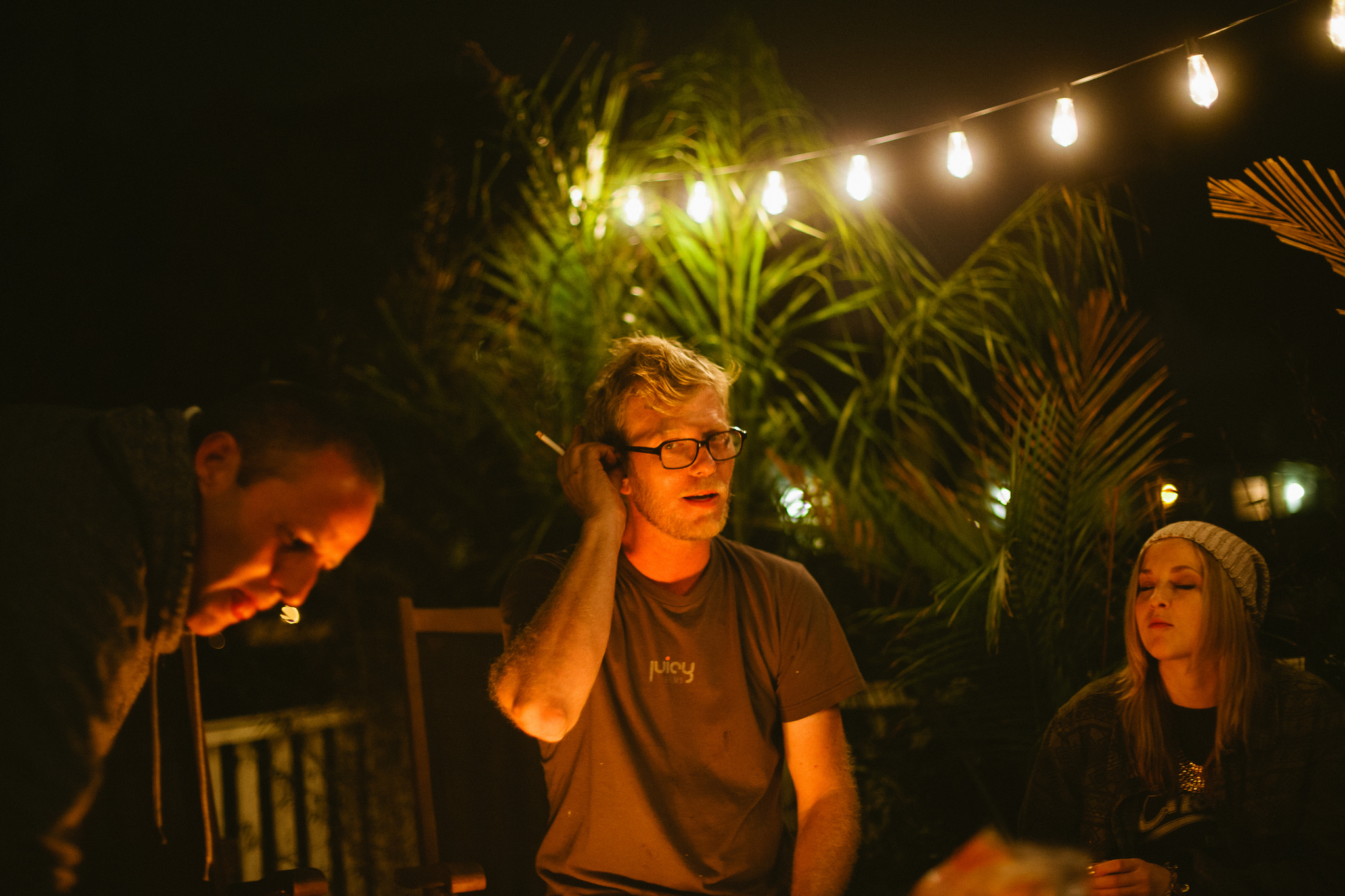 Friends gathered around a backyard fire pit beneath warm string lights with a man in glasses adjusting his hair.