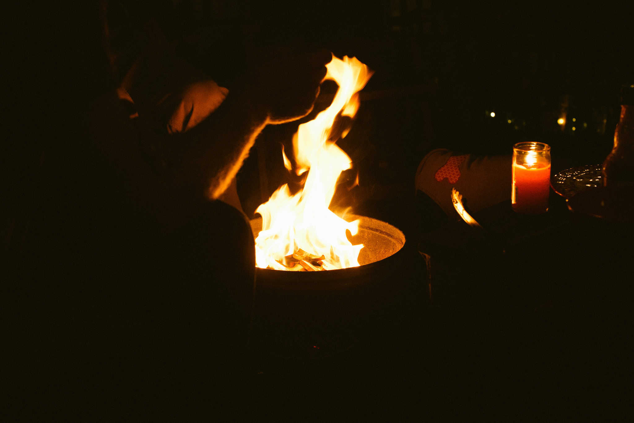 Flames dancing from an outdoor fire pit at night as a person warms their hands beside a glowing candle.