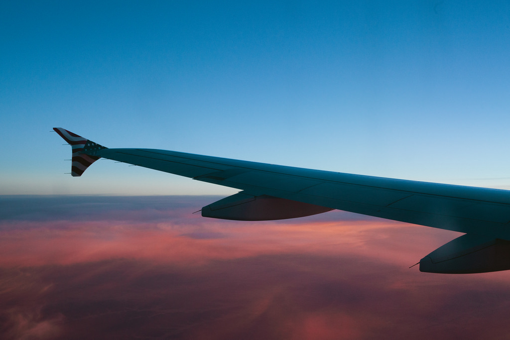 Airplane wing with an American flag winglet soaring above glowing pink and purple sunset clouds against a blue sky.