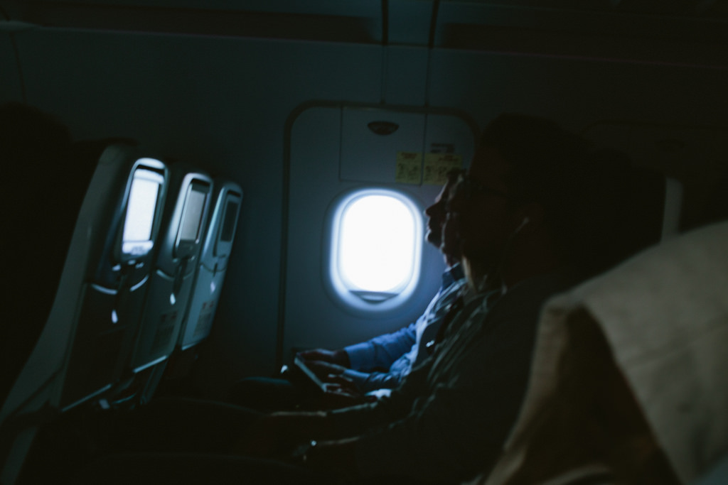 Dimly lit airplane cabin with seated passengers silhouetted against a brightly glowing oval window.