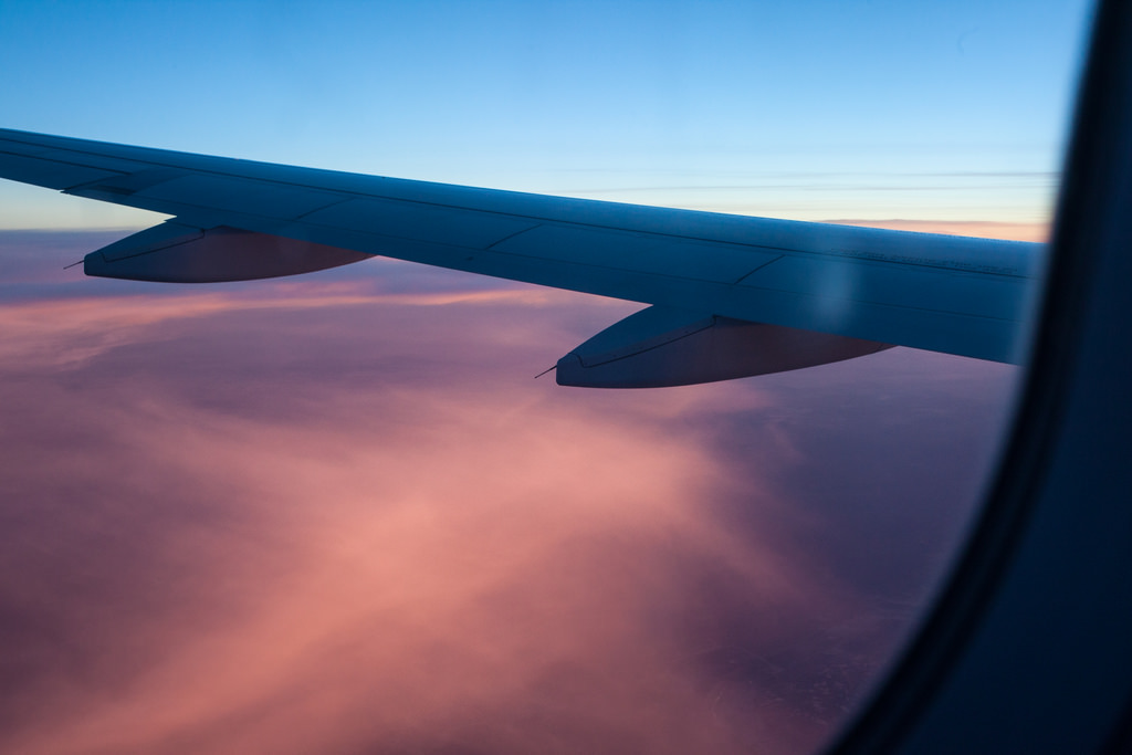 Framed airplane window view of a jetliner wing extending over soft pink and mauve cloud layers at dusk.