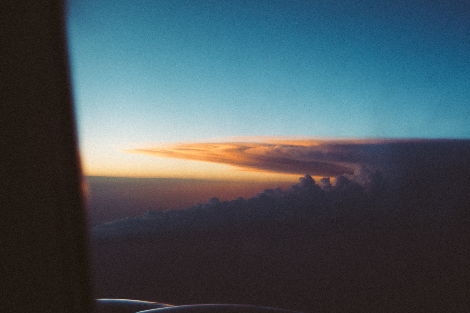 Airplane window view at sunset showing a dramatic anvil cloud formation glowing orange above dusk clouds.
