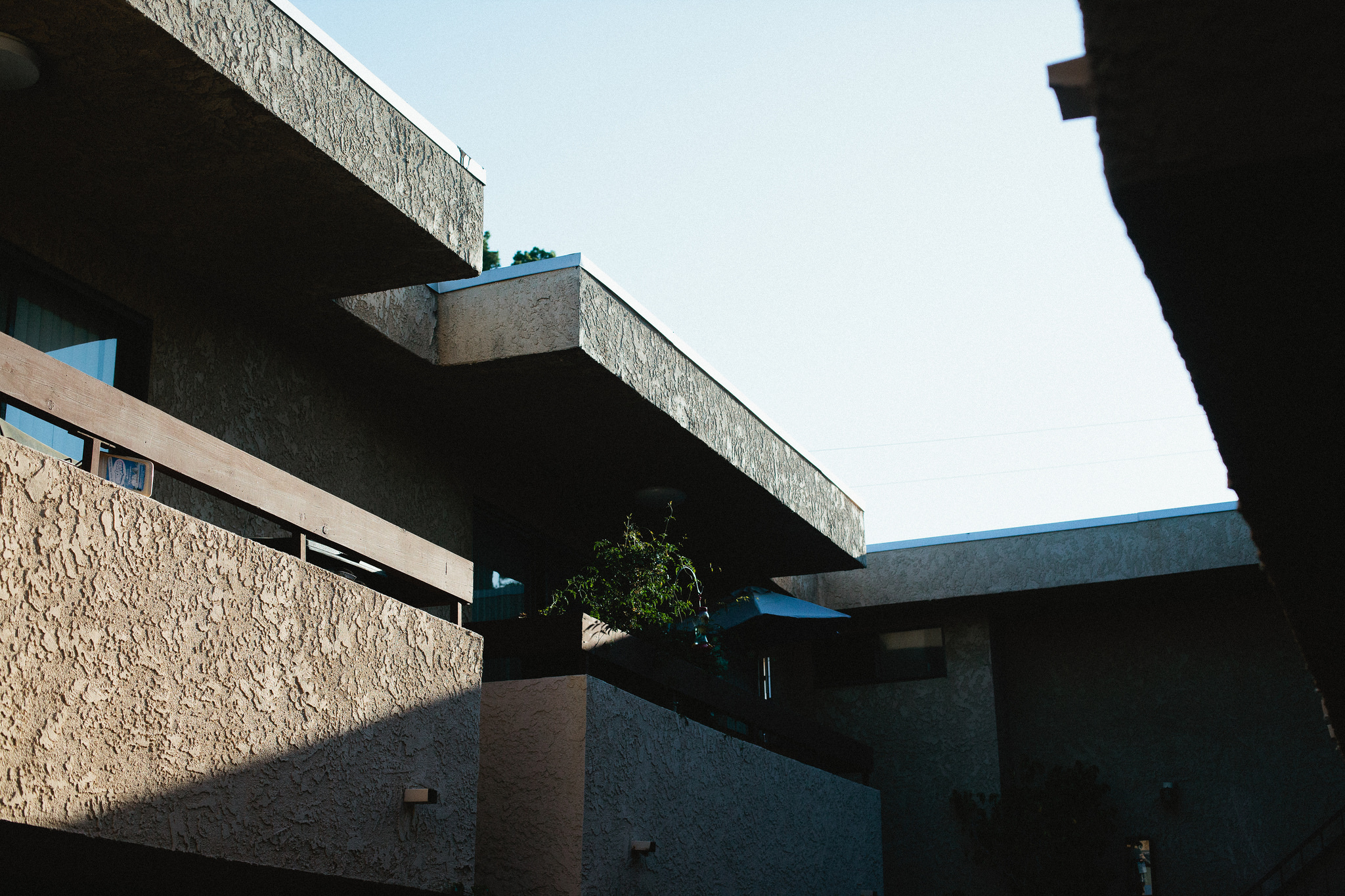 Stucco apartment balconies with geometric diagonal shadows and a potted plant against a clear sky.