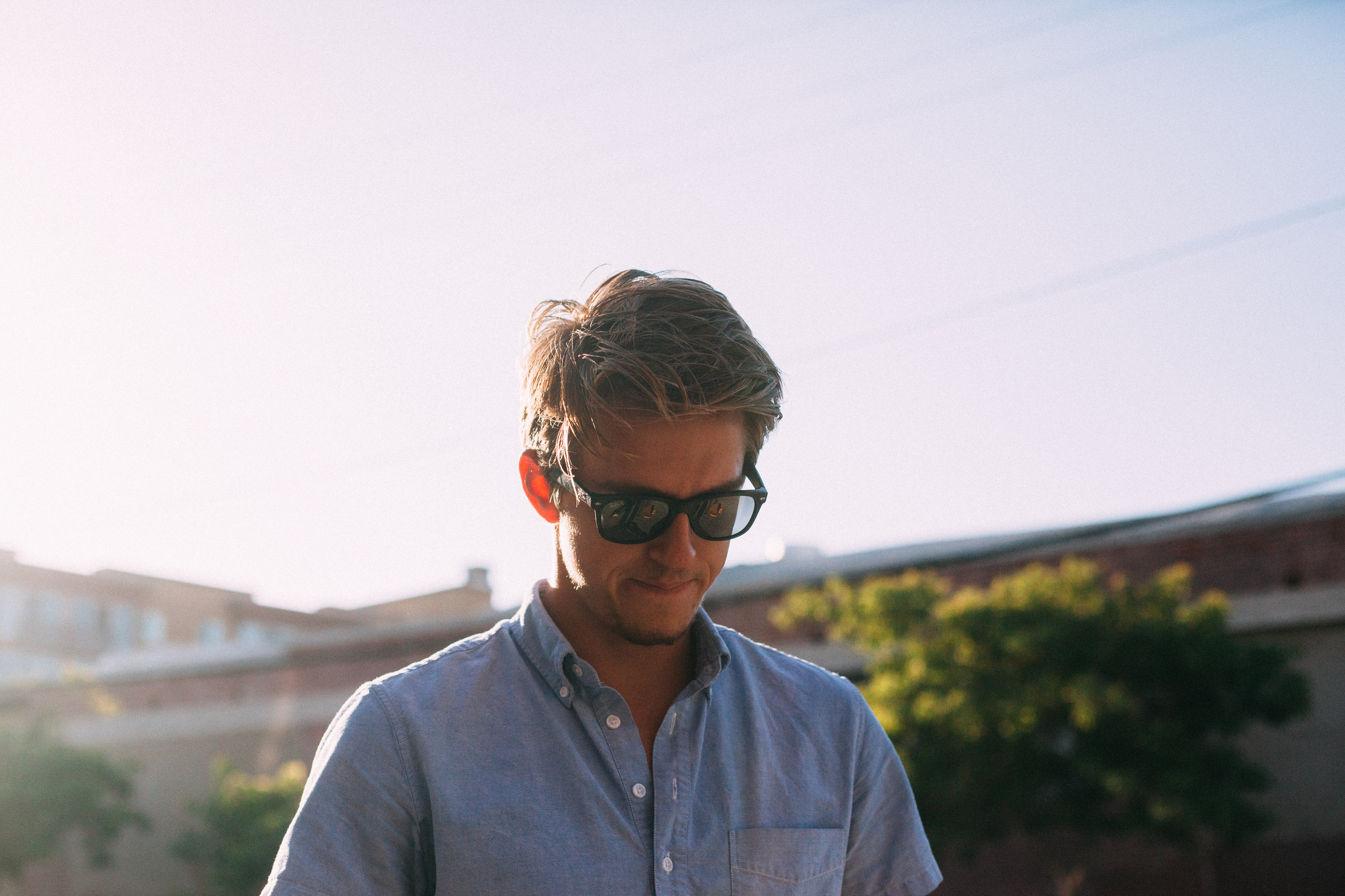Sun-drenched portrait of a young man with tousled hair wearing sunglasses and an unbuttoned blue collared shirt under bright summer sunlight, shot with a Canon 5D Mark II.