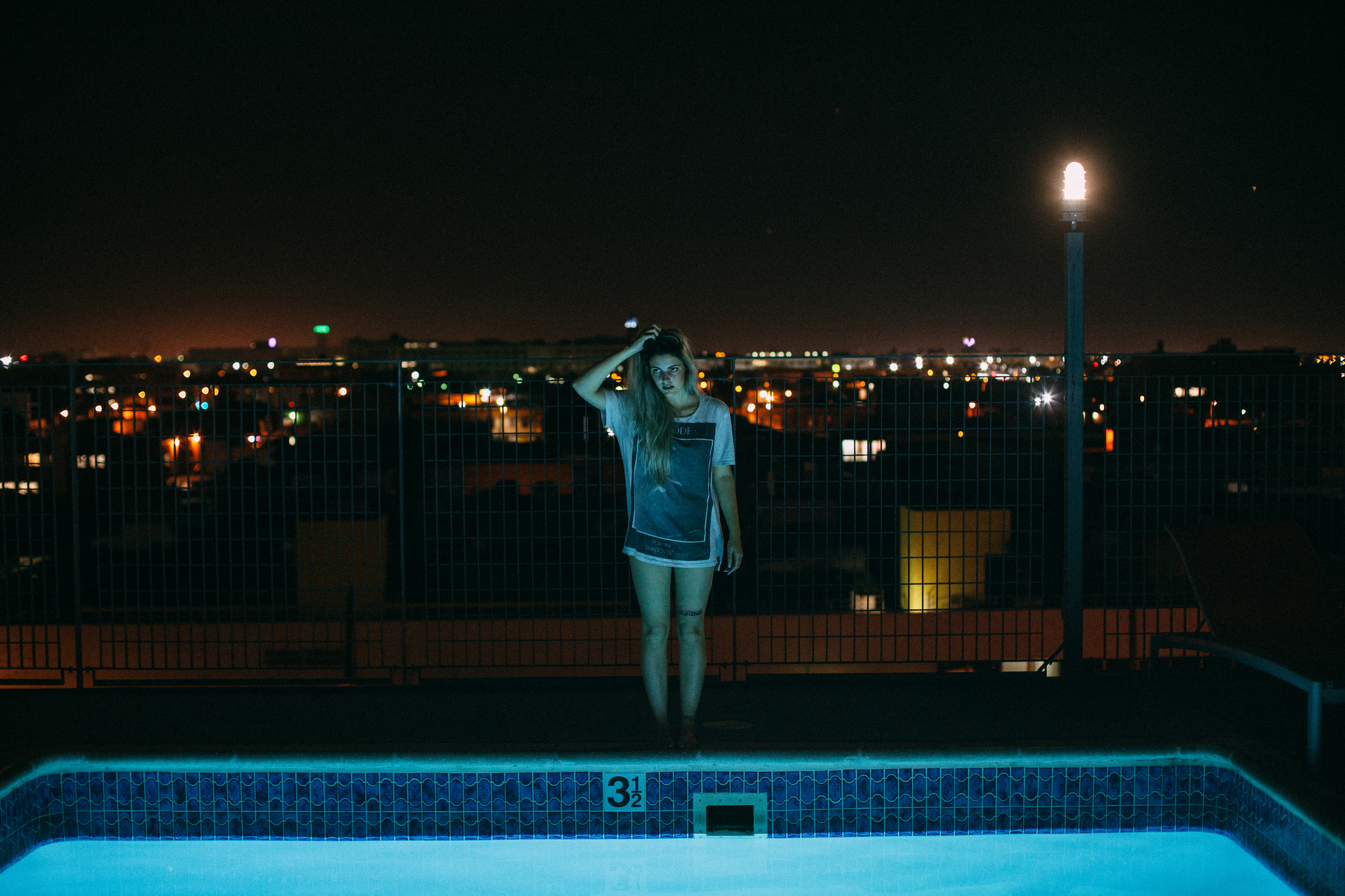 Shelby Parks touching her hair beside an illuminated turquoise pool at night against a city skyline.