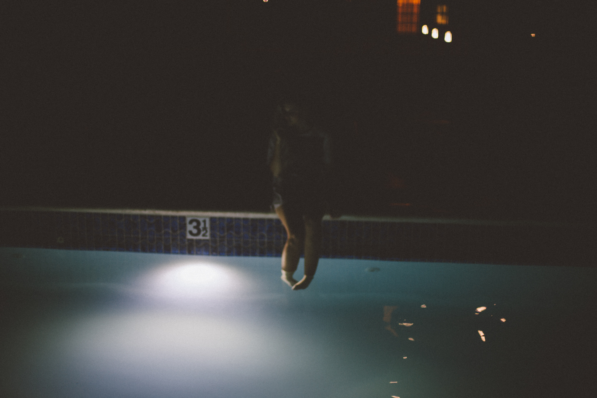 Shelby Parks sitting on a pool ledge at night dangling her legs in the water beside a 3 1/2 foot marker.