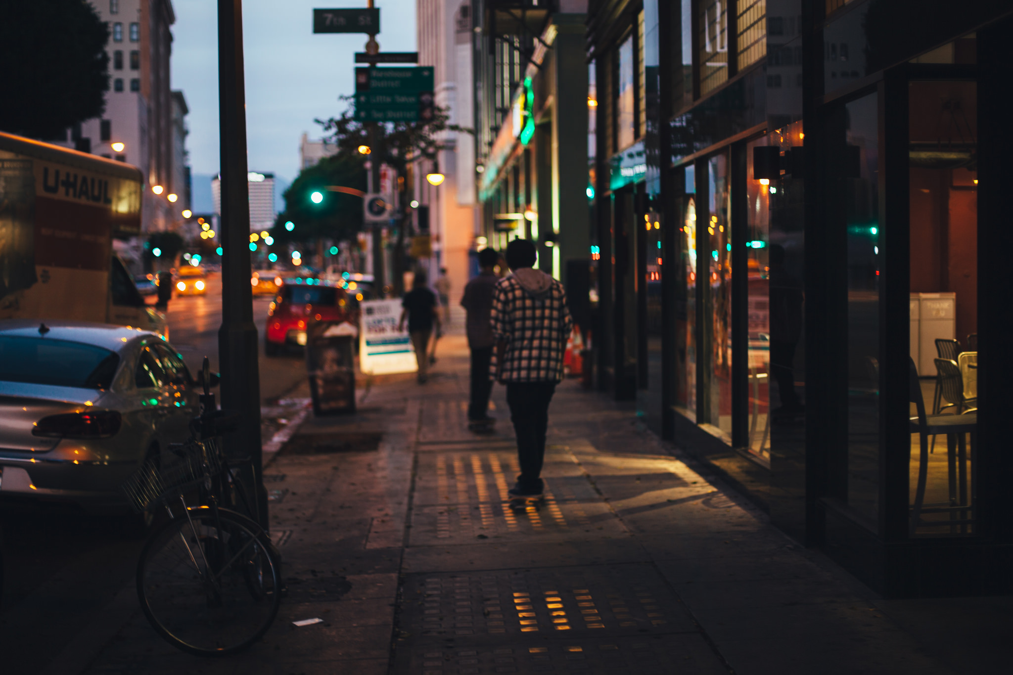 Pedestrians on a dusk city sidewalk on 7th Street with glowing streetlamps and storefront lights.