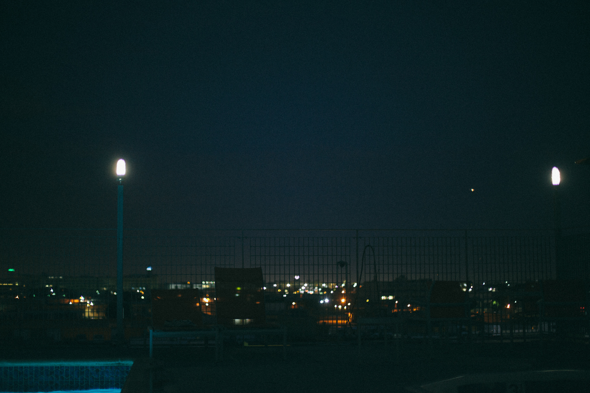 Nighttime view from a rooftop terrace overlooking a wire fence with glowing post lamps and twinkling distant city lights, shot with a Canon 5D Mark II.