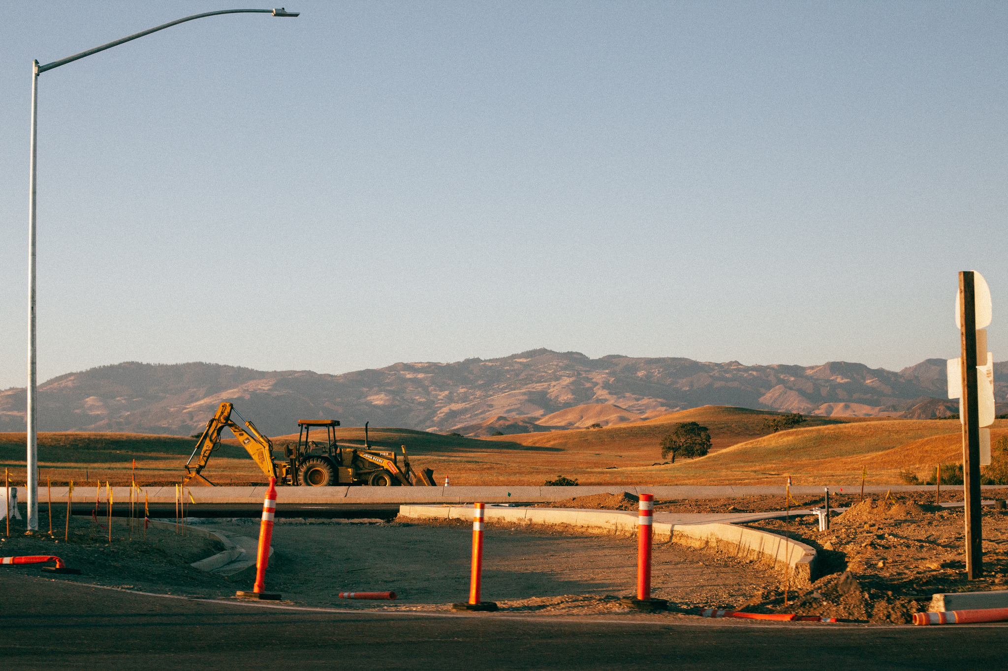 Yellow backhoe parked on a graded road shoulder with orange traffic cones and rolling hills behind it.
