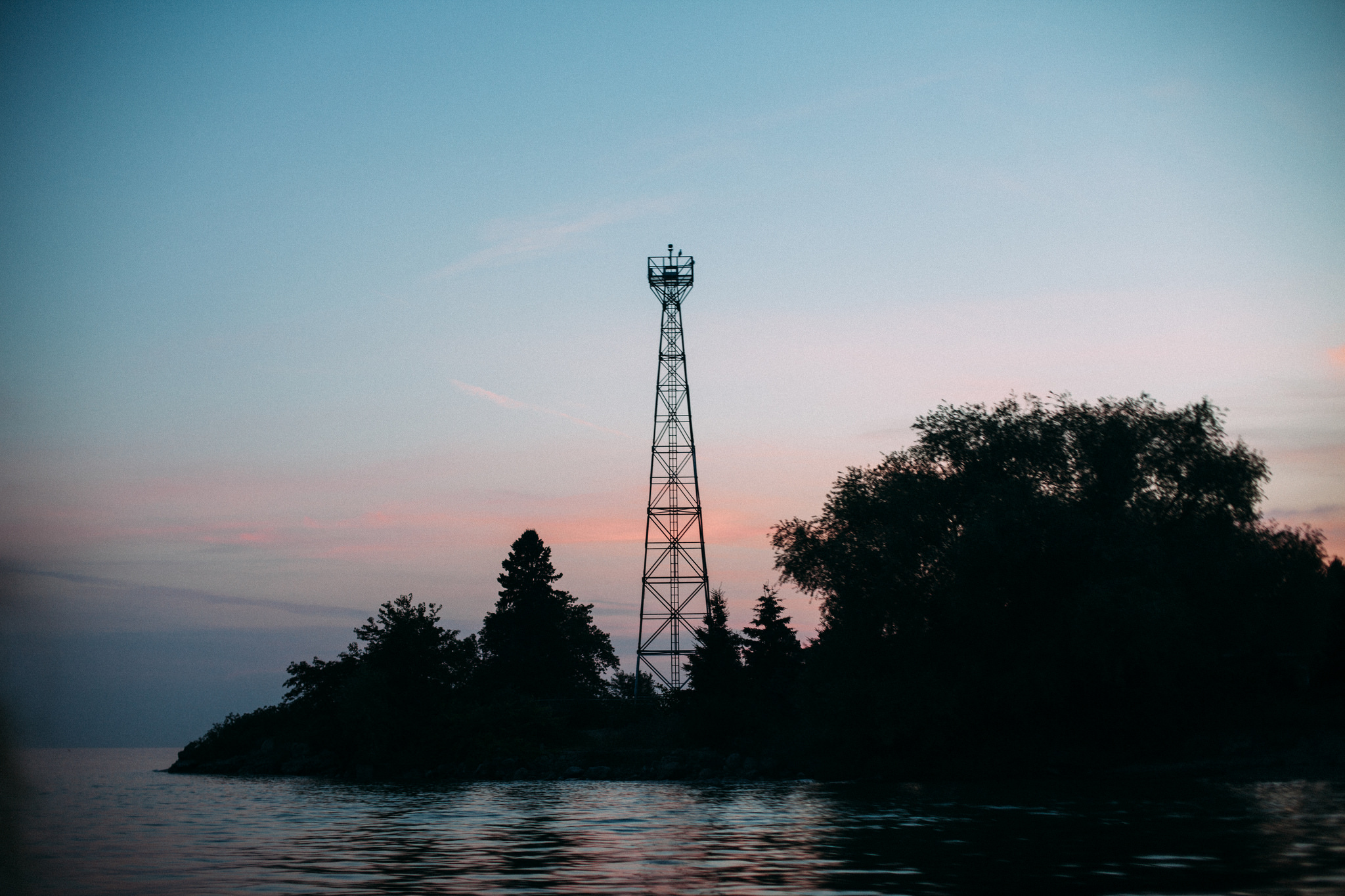 Silhouette of a steel observation tower rising above pine trees on a rocky island shoreline at dusk.