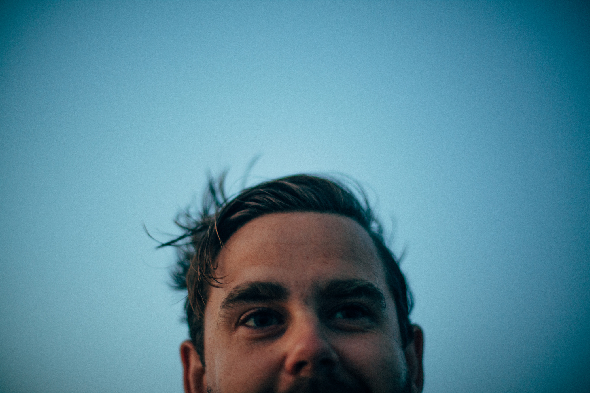 Close-up portrait of the upper face and eyes of a man with windblown hair looking upwards against a blue sky.