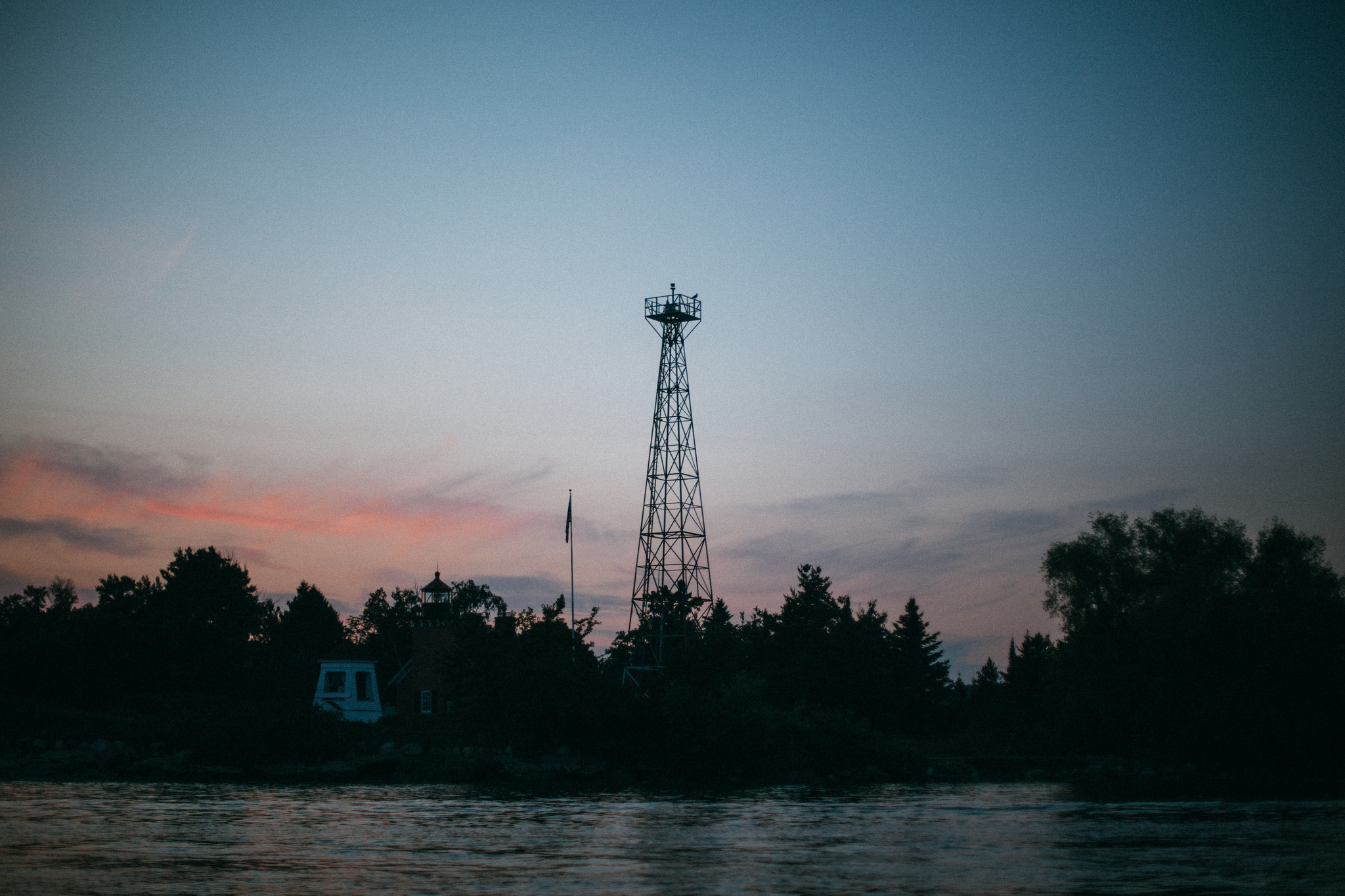 Steel beacon tower and lighthouse silhouetted against a twilight sky with pink sunset clouds over water.