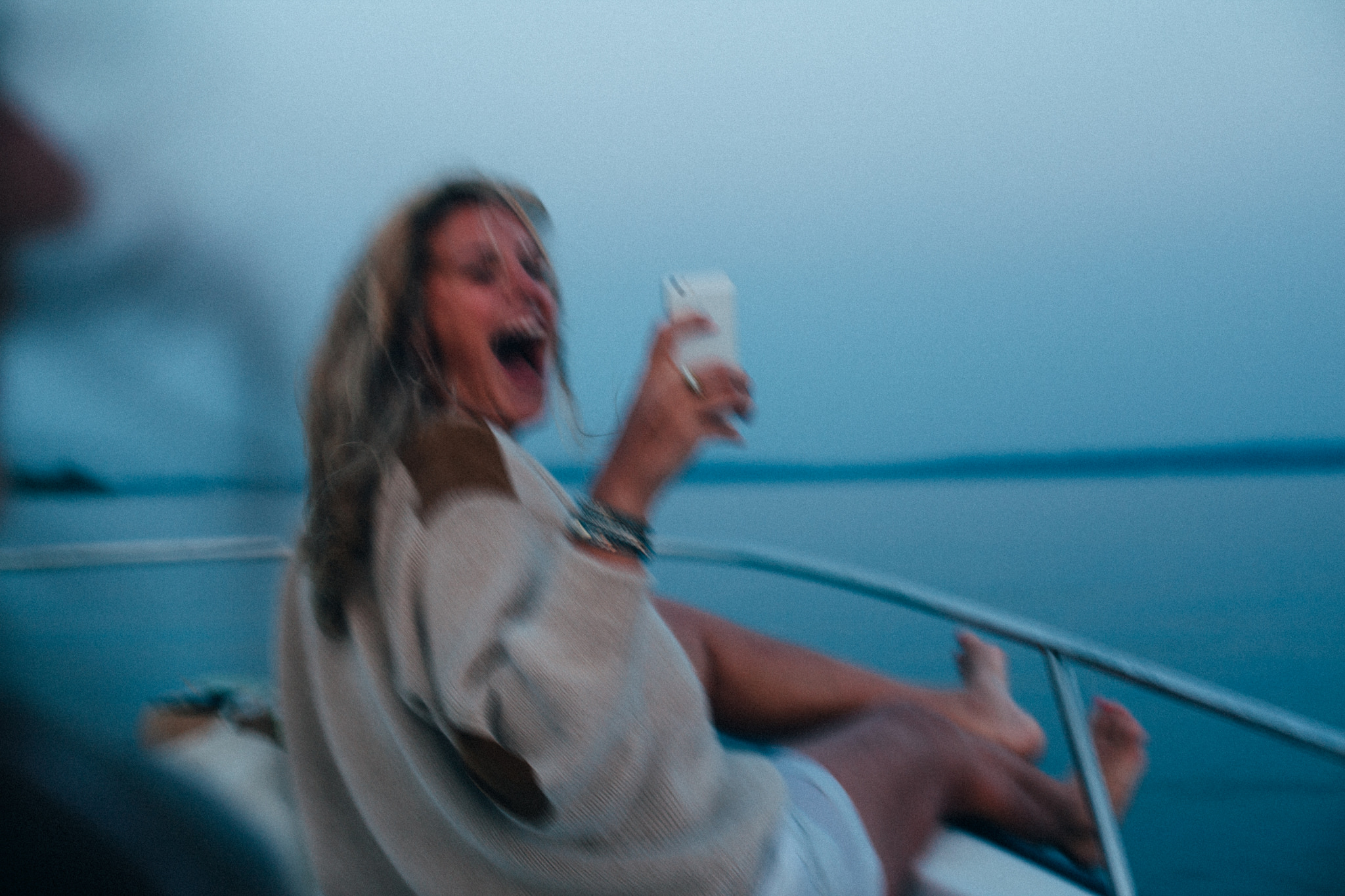 Laughing woman holding up a white smartphone on the bow of a moving boat at dusk.