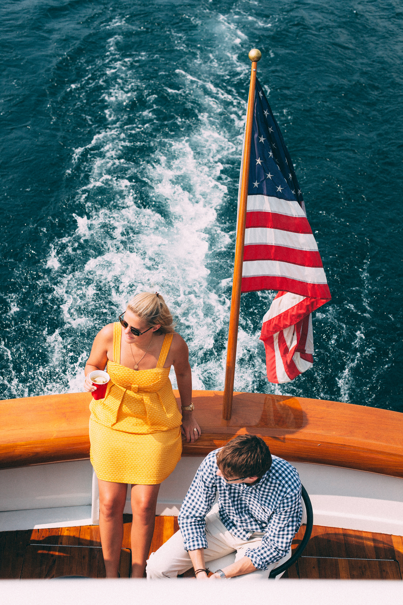 High-angle view of a woman in a yellow sundress and a man in a blue gingham shirt on the wooden deck of a boat flying an American flag over churning water, shot with a Canon 5D Mark II.