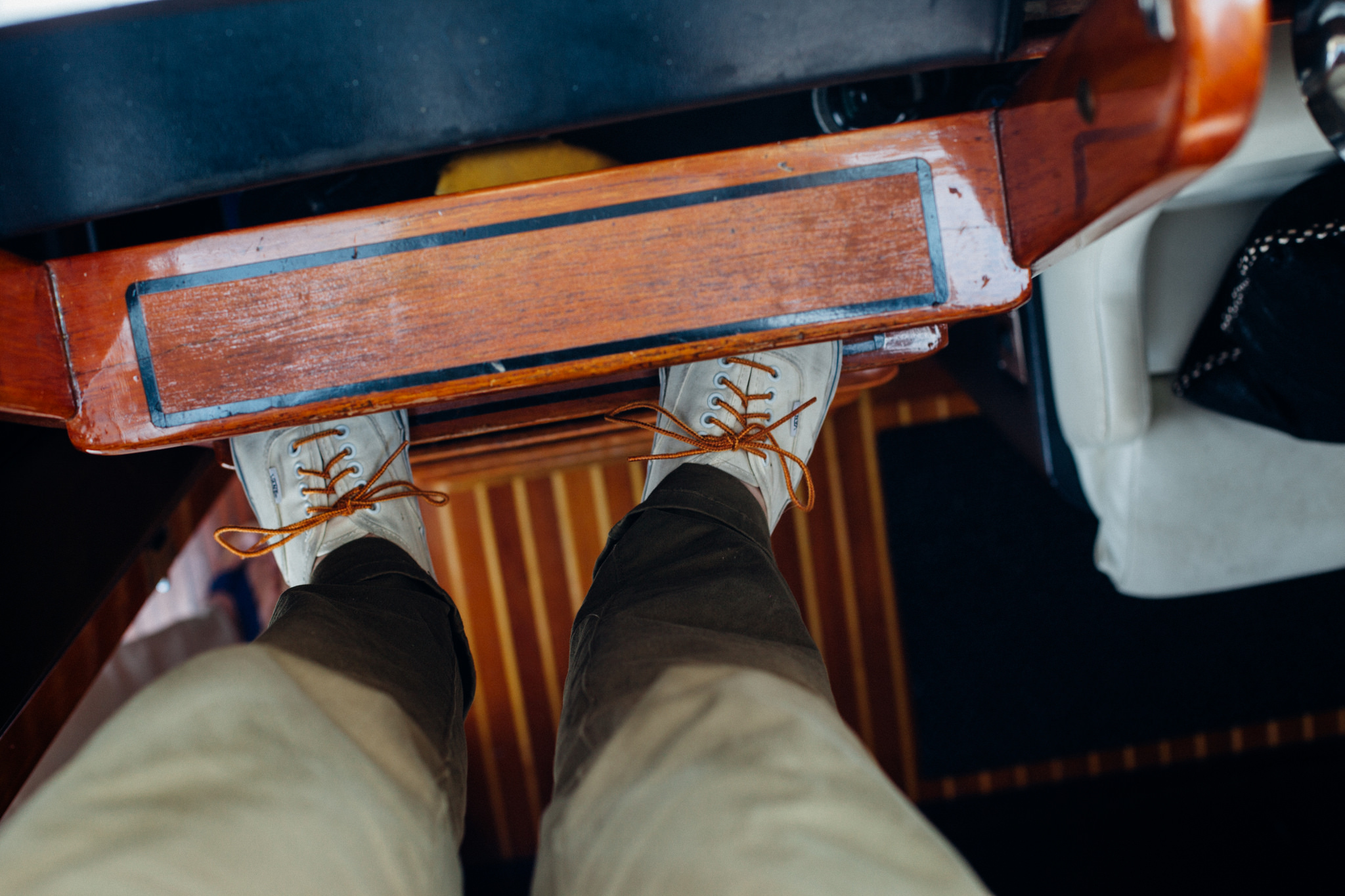 First-person perspective looking down at white canvas lace-up sneakers resting on a varnished mahogany step above striped boat flooring, shot with a Canon 5D Mark II.