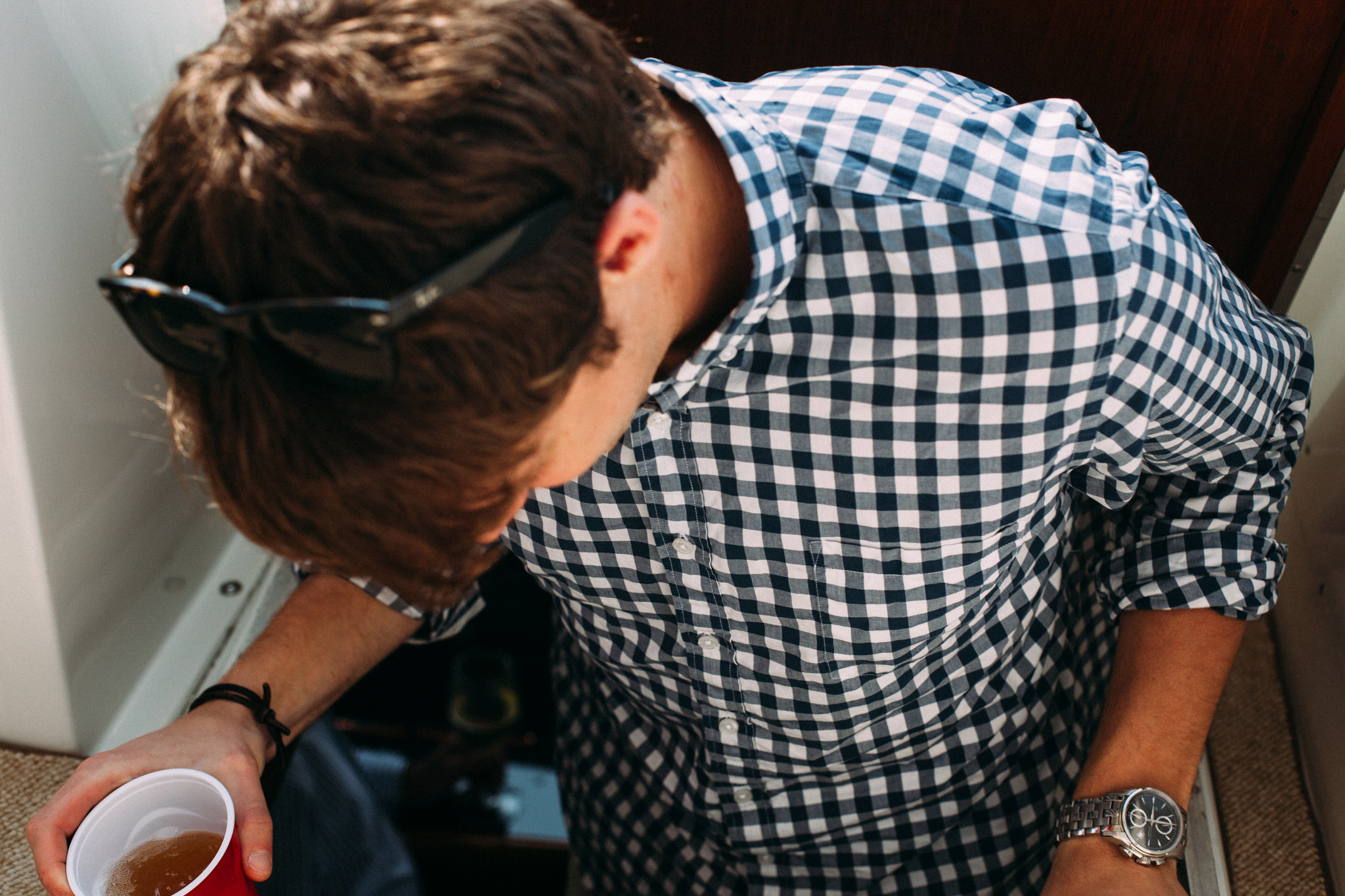 Overhead close-up of a man in a blue and white gingham check shirt holding a cup of beer on a boat deck, shot with a Canon 5D Mark II.