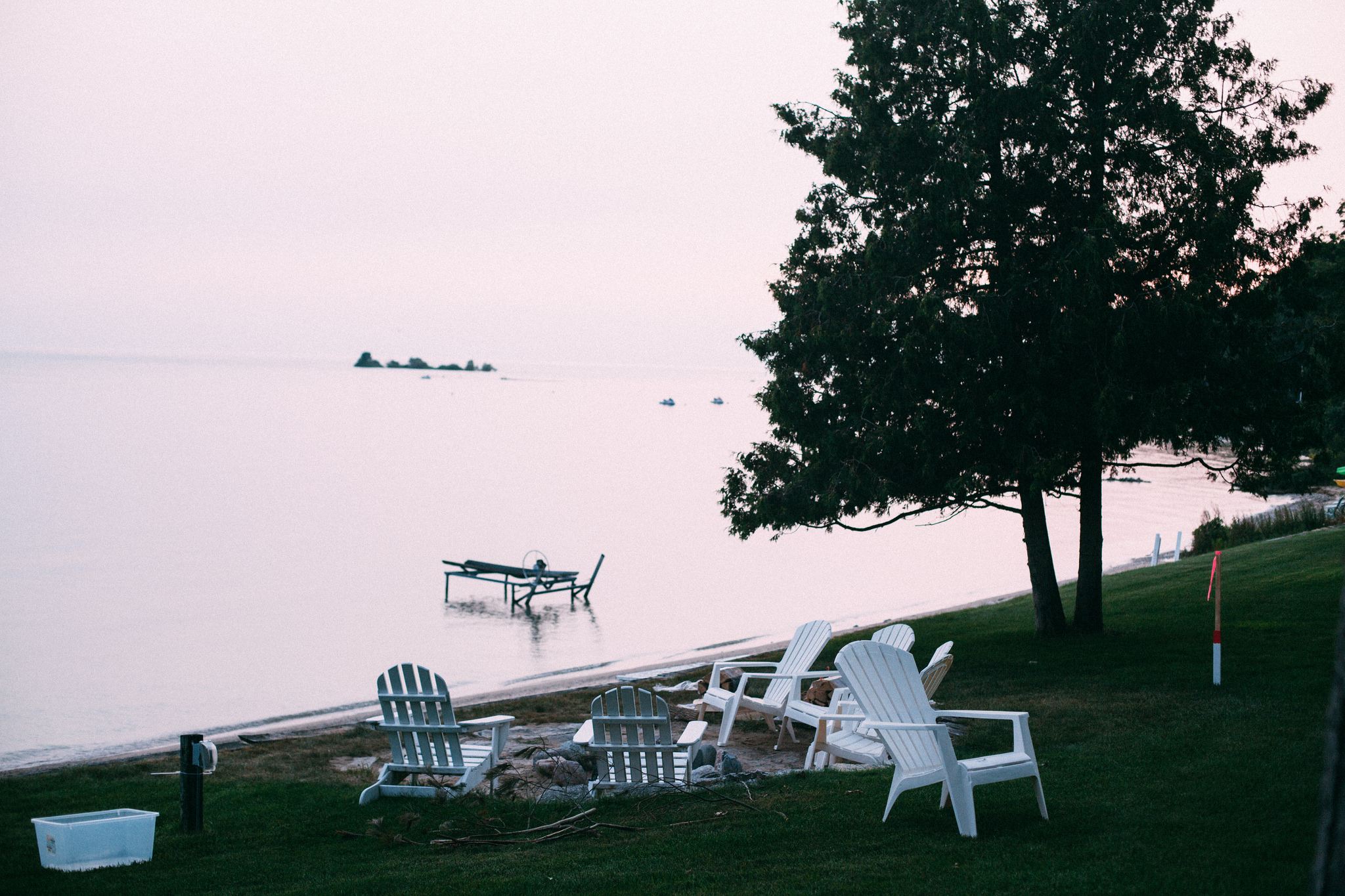 White Adirondack chairs around a fire pit on a lakeside lawn beneath pine trees overlooking calm water.