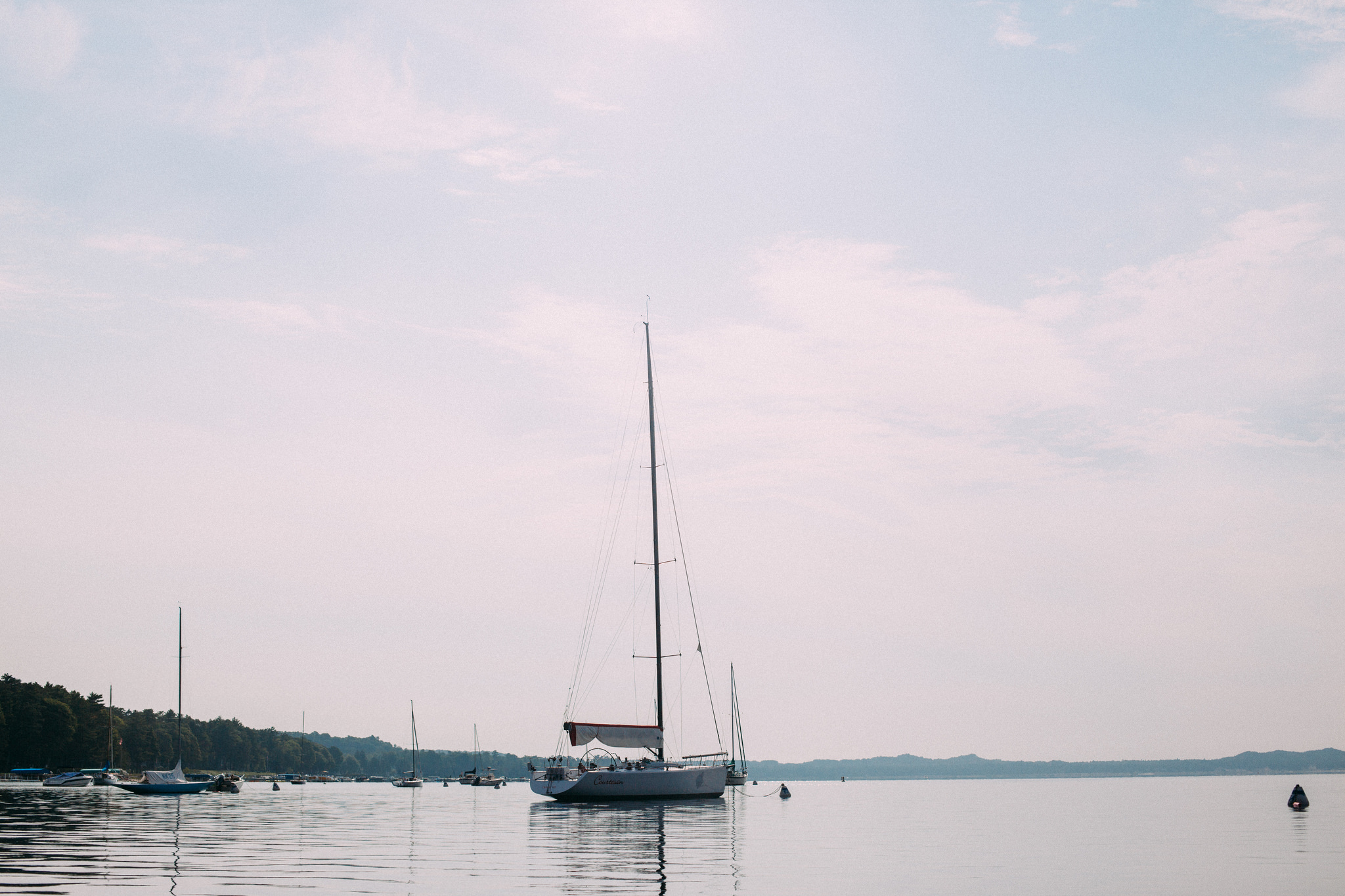 A white sailboat at anchor on the calm, flat waters of Lake Michigan under a soft hazy evening sky.