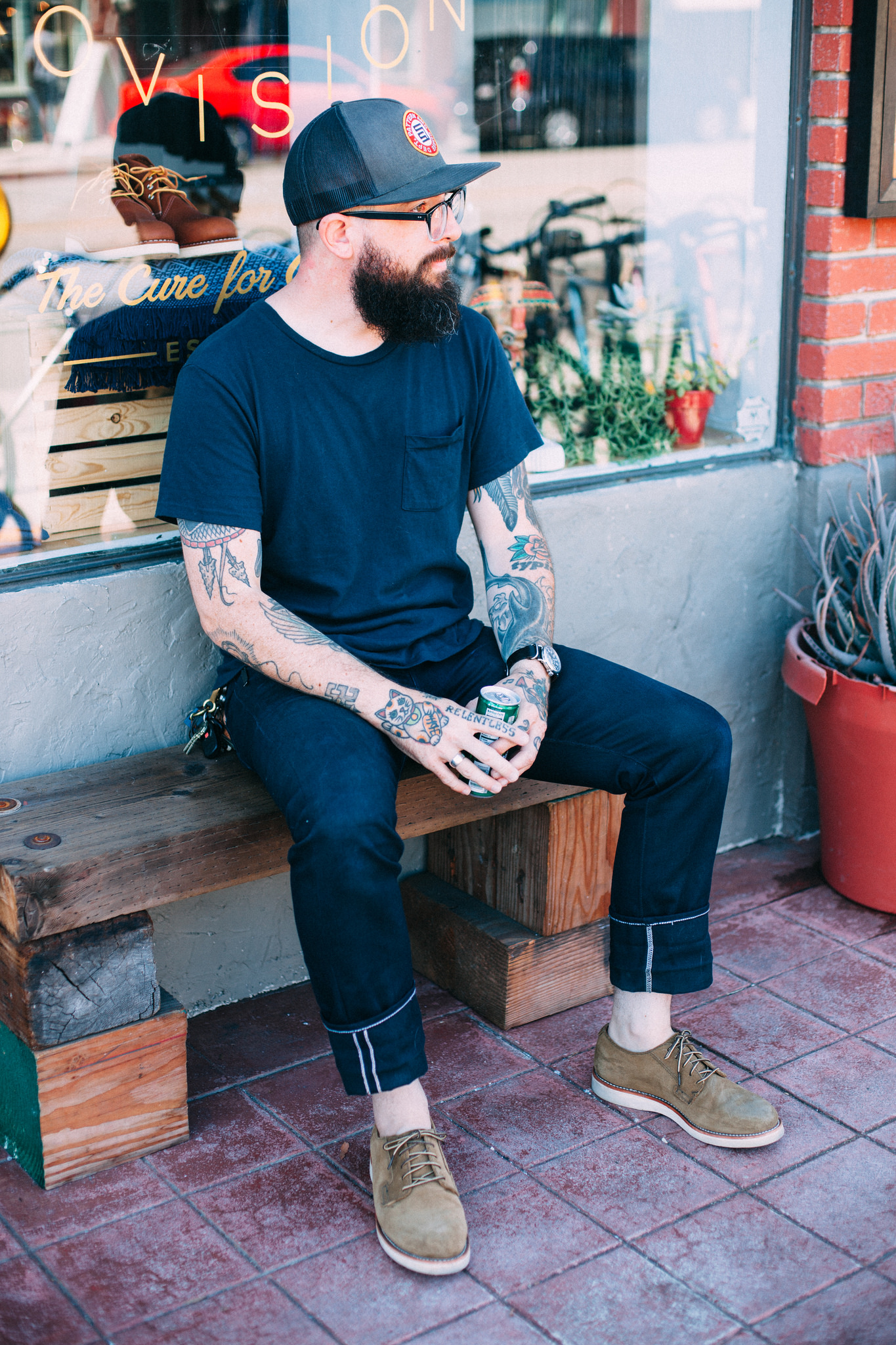 Full-length seated portrait of a bearded man in a trucker cap, cuffed dark denim jeans, and tan suede shoes on a wooden bench outside Snake Oil Provisions, shot with a Canon 5D Mark II.