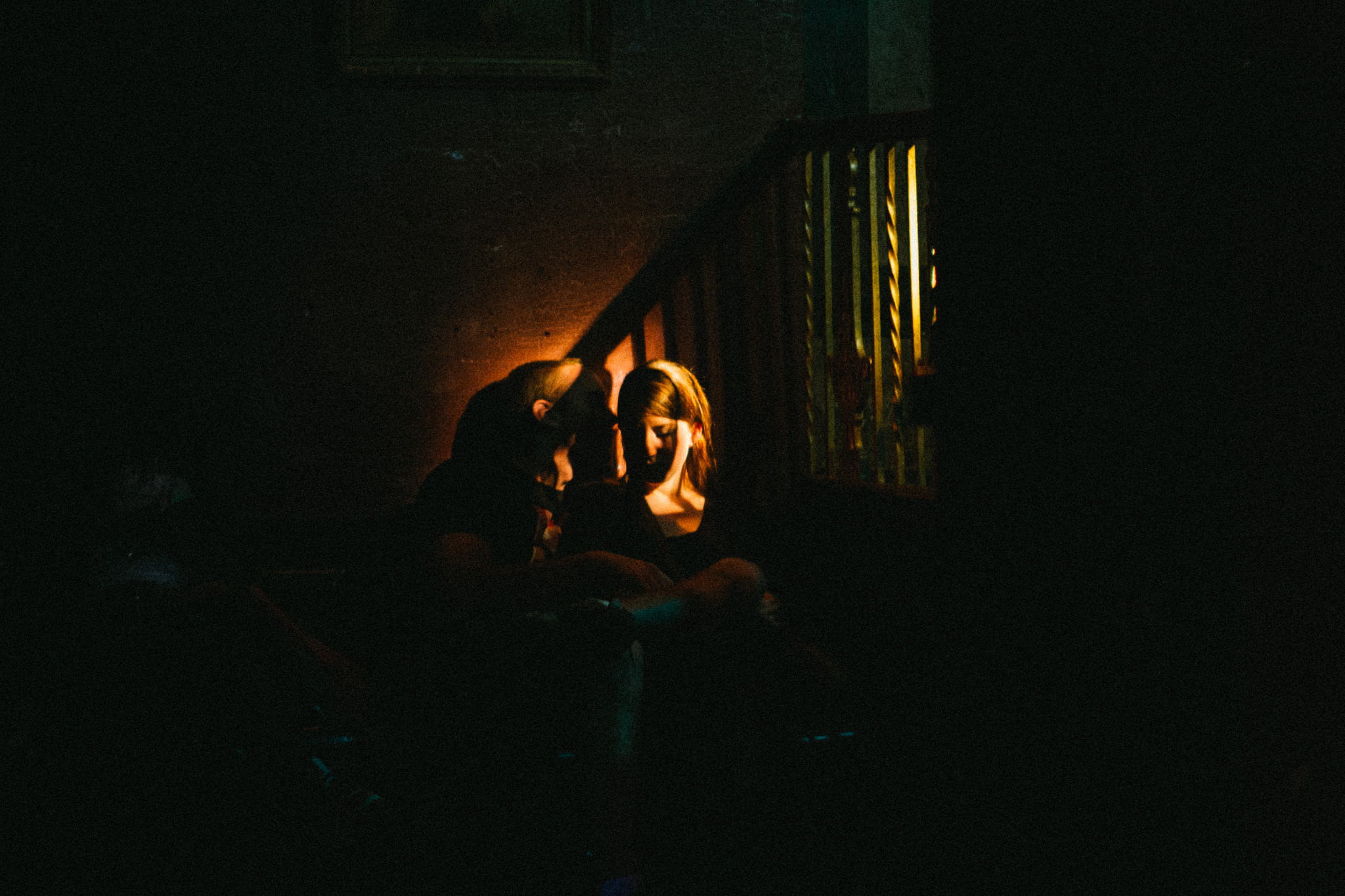 Couple seated by a wooden staircase in a dim interior, illuminated by a spotlight with dramatic shadows.