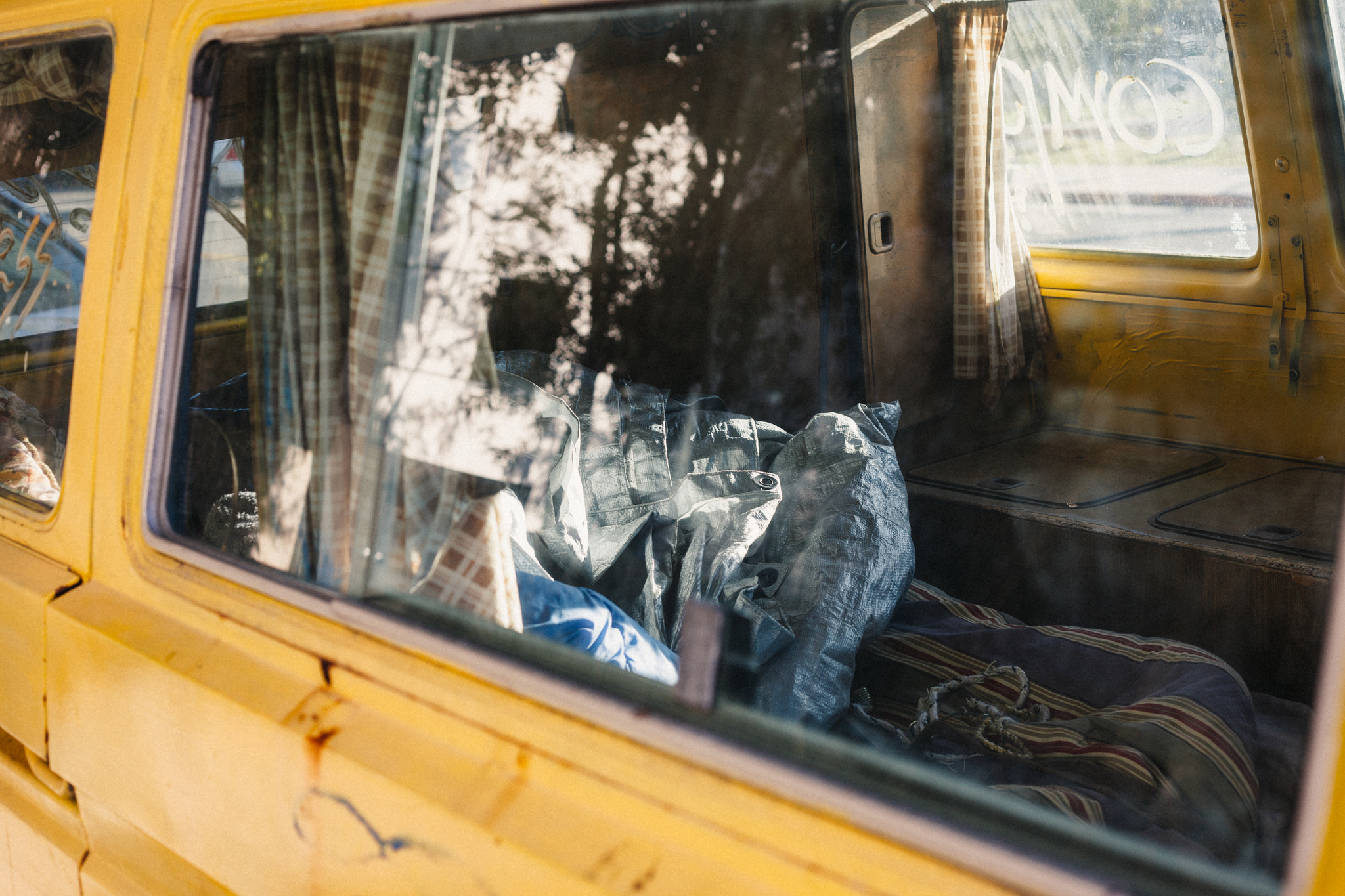 Yellow camper van interior viewed through a window, showing plaid curtains, folded blankets, and a bed.