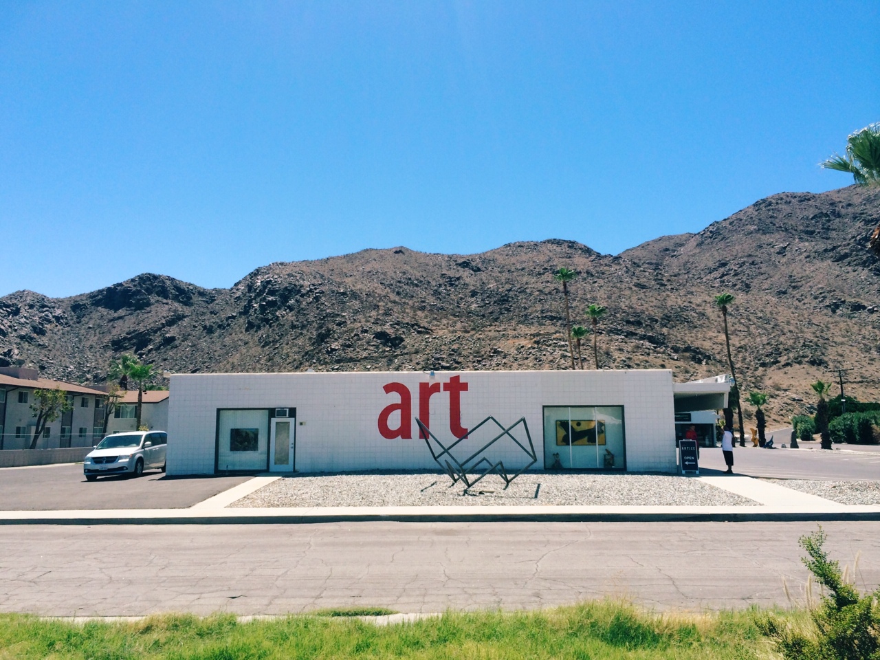 White gallery displaying red 'art' signage with a metal sculpture before a rocky desert mountain.