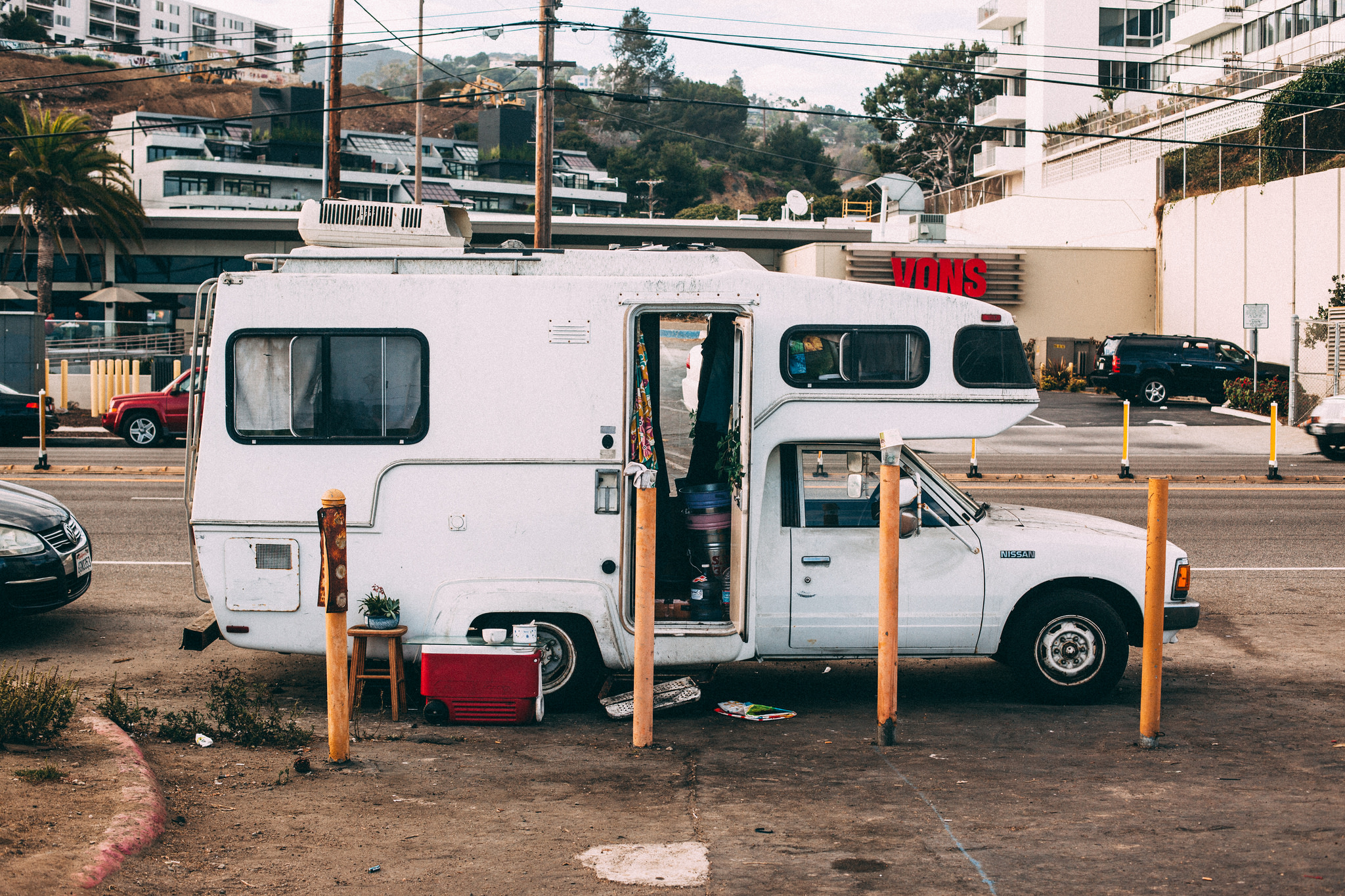 A vintage white Nissan mini-motorhome camper parked on a paved lot with its side door open showing living quarters, with yellow bollards and a Vons grocery store in the background, shot with a Canon 5D Mark II.