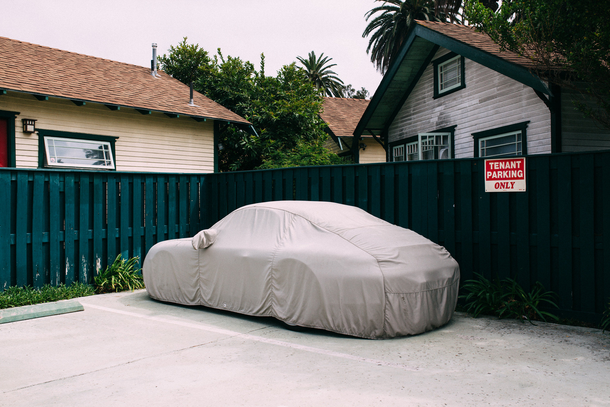 Sports car under a beige protective car cover parked in a stall beside a green wooden fence and house.