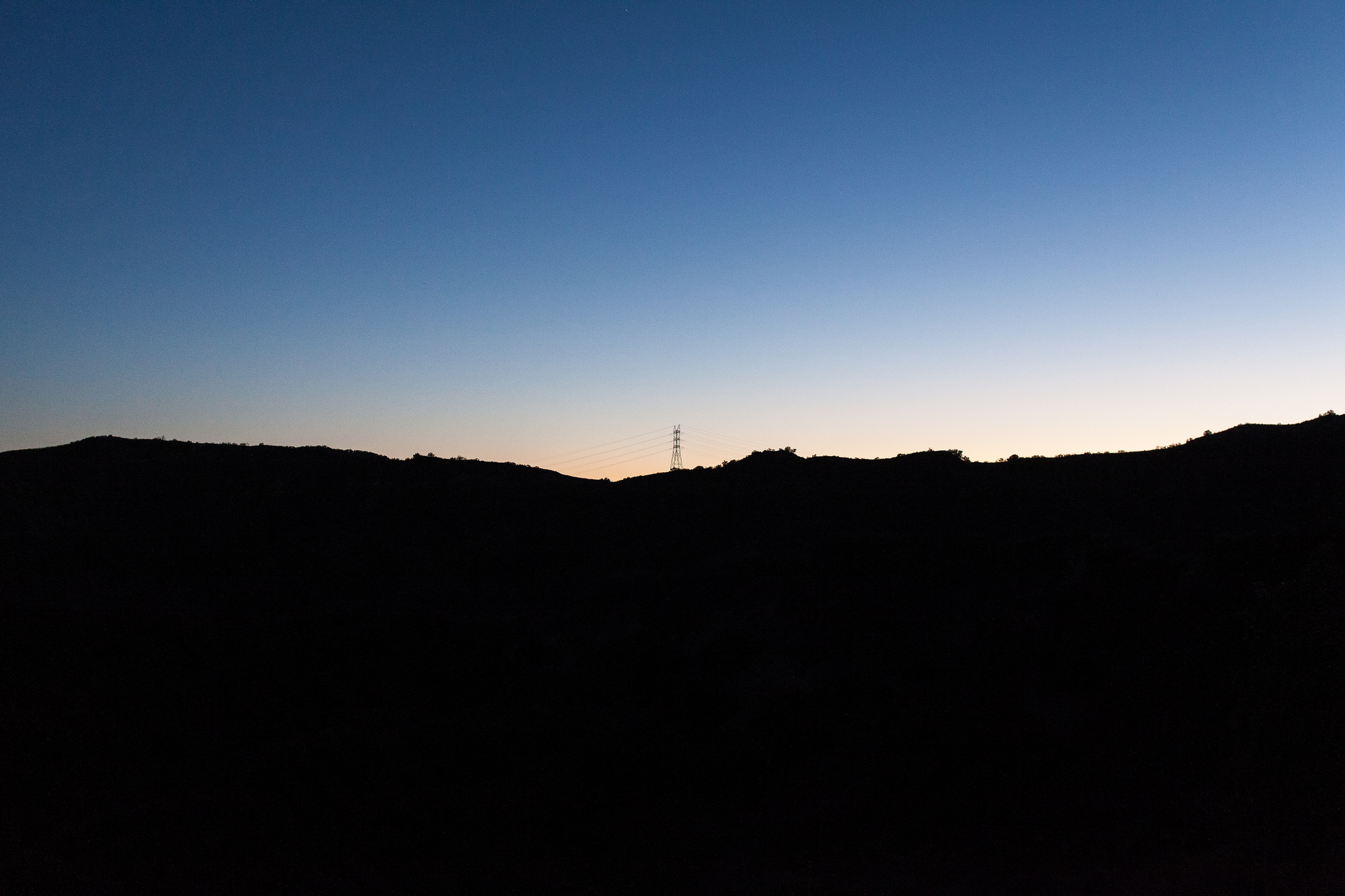 Dark silhouette of rolling hills and a transmission tower under a deep blue and orange twilight sky.
