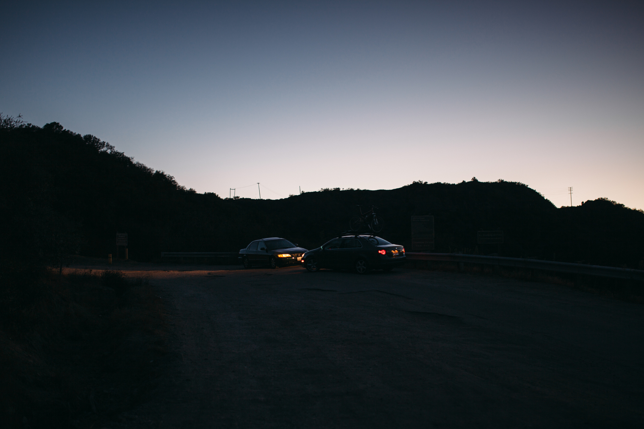 Two cars with taillights illuminated parked at a mountain overlook at dusk against dark hill silhouettes.