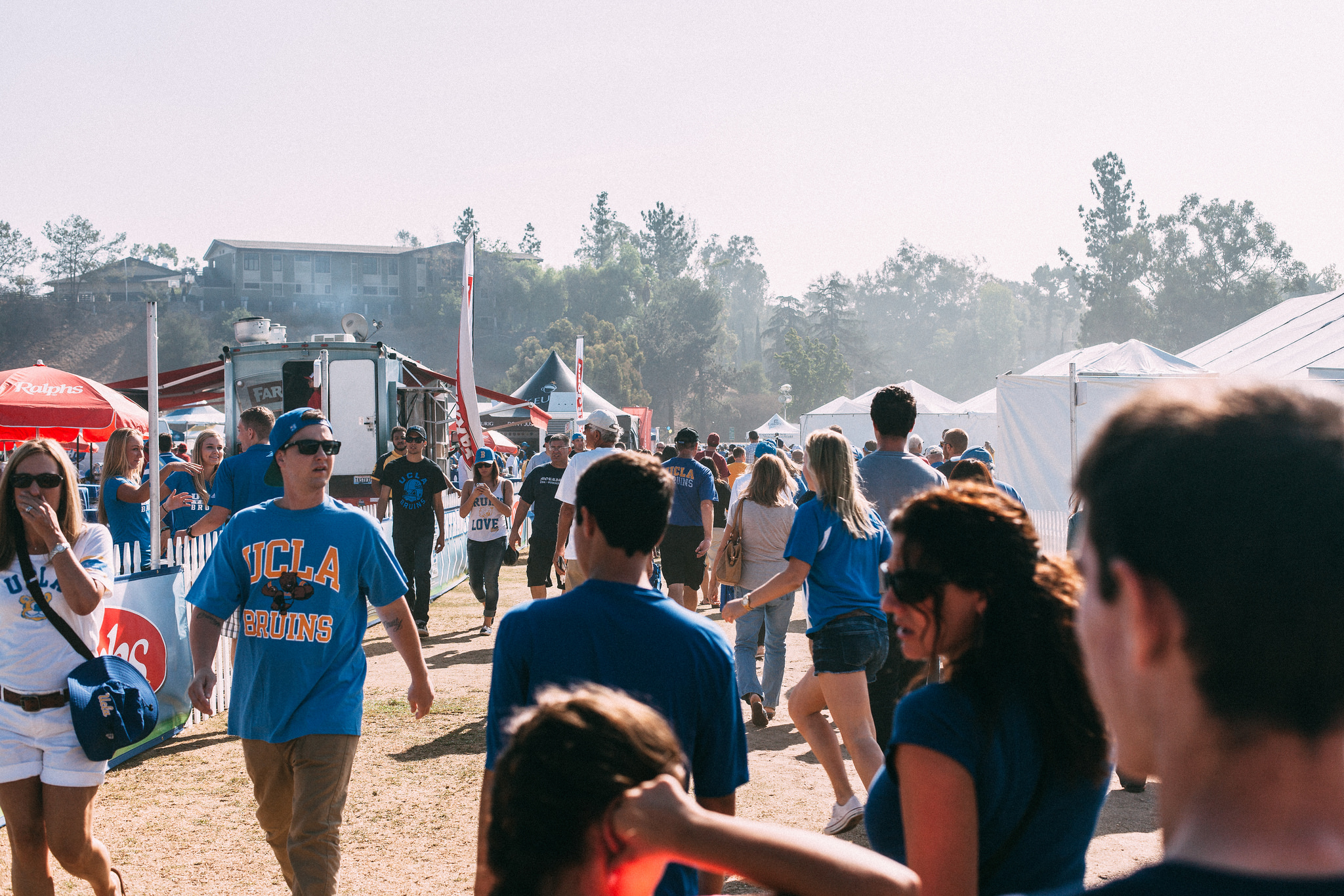 Football fans in UCLA blue t-shirts walking through a bustling tailgate festival outside the stadium.