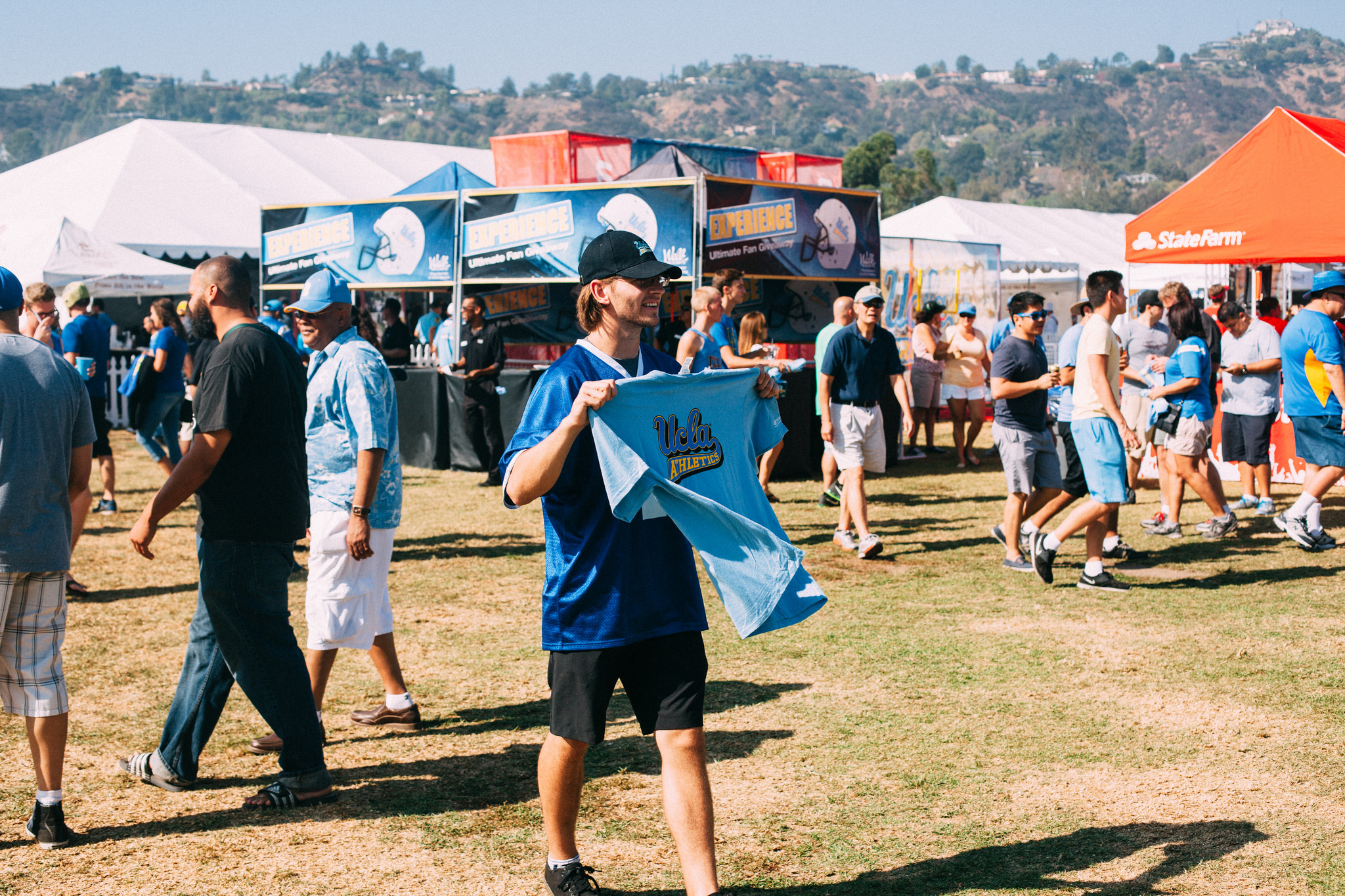 Fan in a jersey holding up a light blue UCLA Athletics t-shirt on the sunny lawn outside the Rose Bowl.