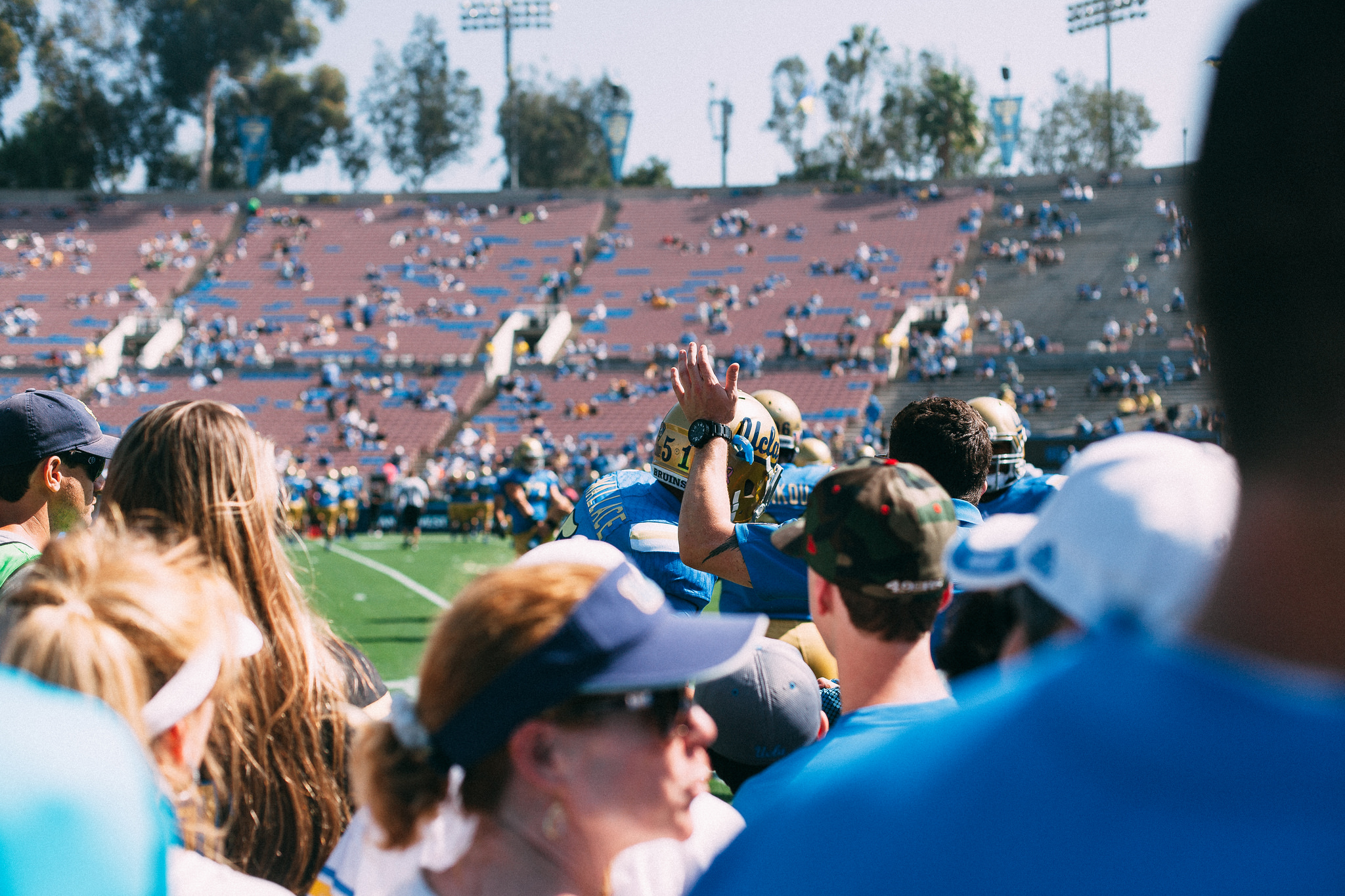 Spectators in the stands watching a UCLA football player in a gold helmet raise his hand on the sideline.
