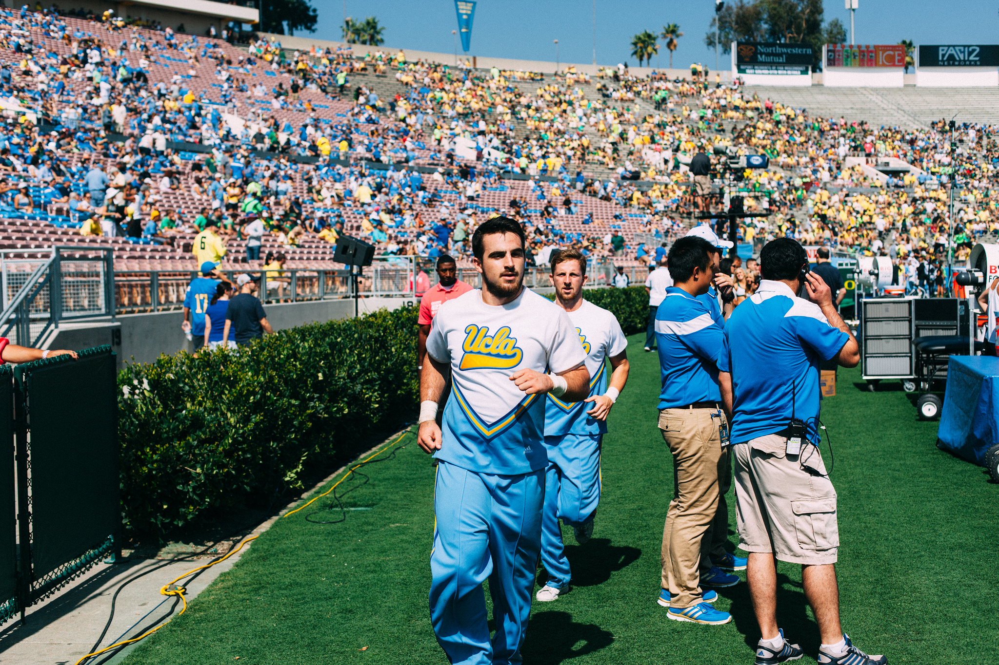 UCLA yell leaders running in light blue uniforms along the sideline before grandstands at the Rose Bowl.