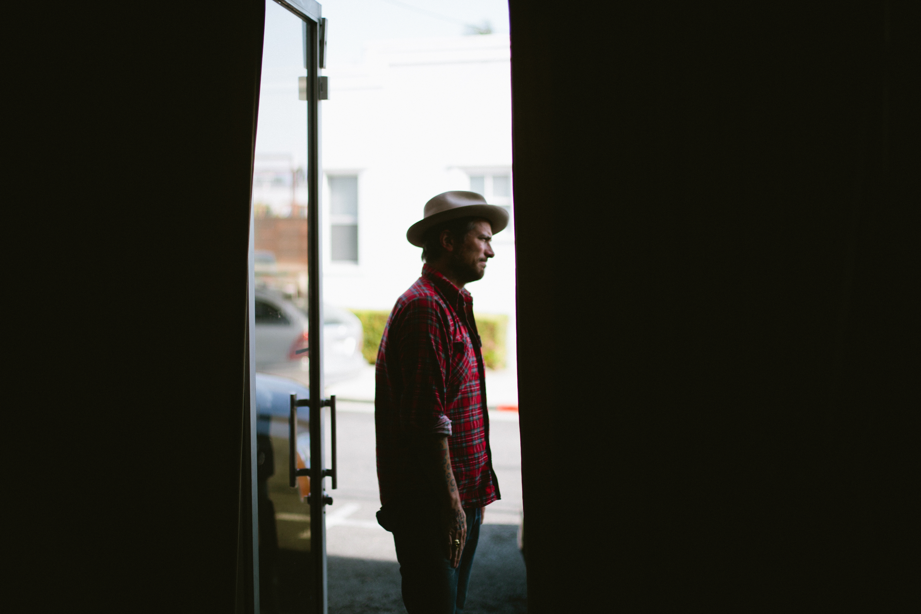 Musician Butch Walker in a wide-brim hat and plaid shirt, standing in a doorway looking into sunlight.
