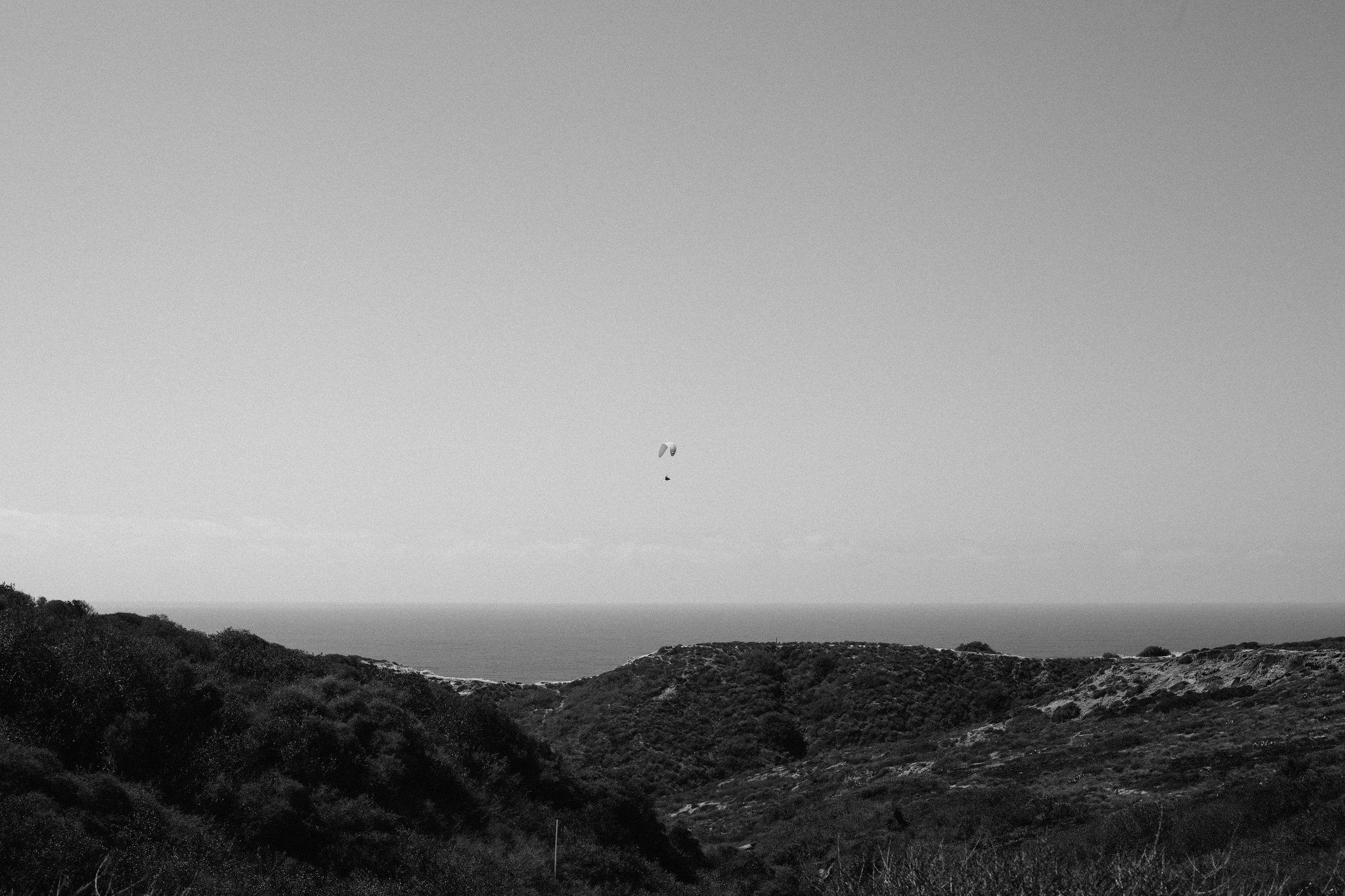 Black and white wide landscape showing a lone paraglider floating high in the sky above coastal hills and the Pacific Ocean, shot with a Canon 5D Mark II.