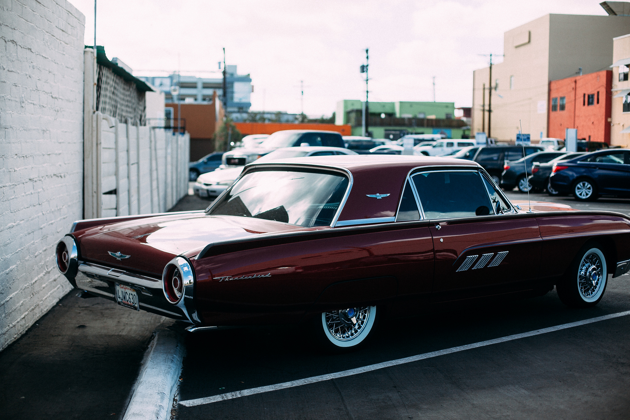 Rear three-quarter view of a vintage maroon Ford Thunderbird coupe parked along a white brick wall.
