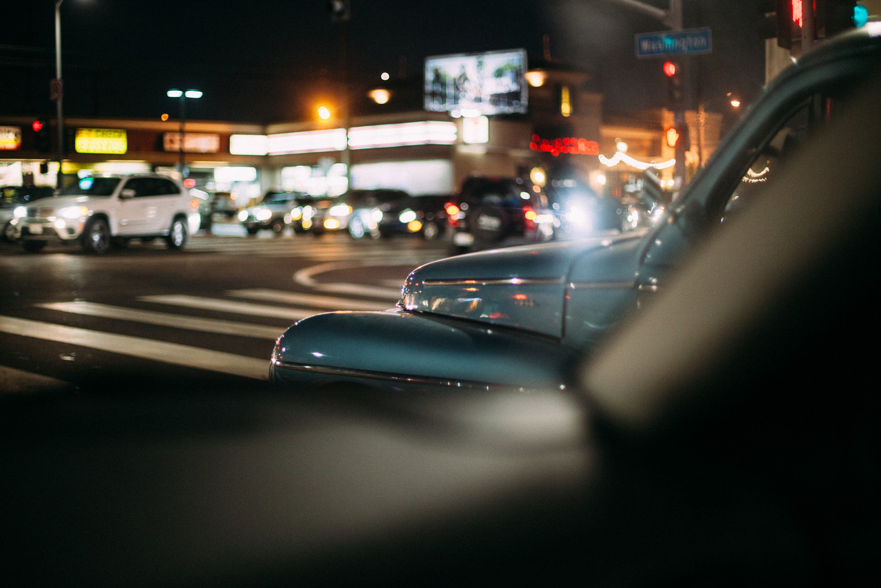 Vintage blue car fender at a city intersection with glowing neon signs and traffic lights at night.