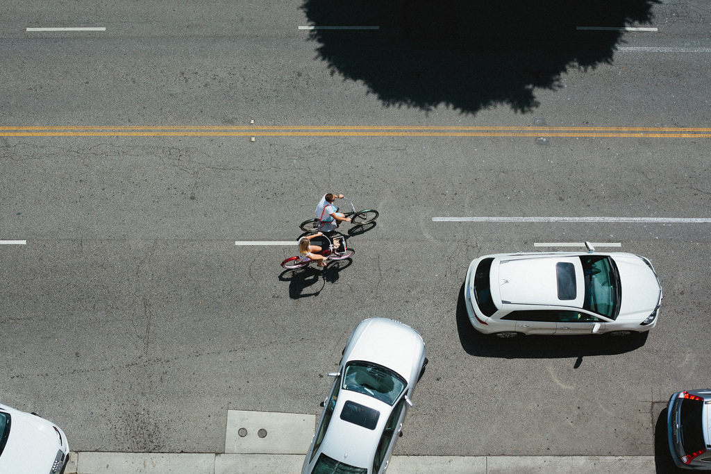 Bird's-eye view of two bicyclists riding past parked and moving cars along an asphalt street.