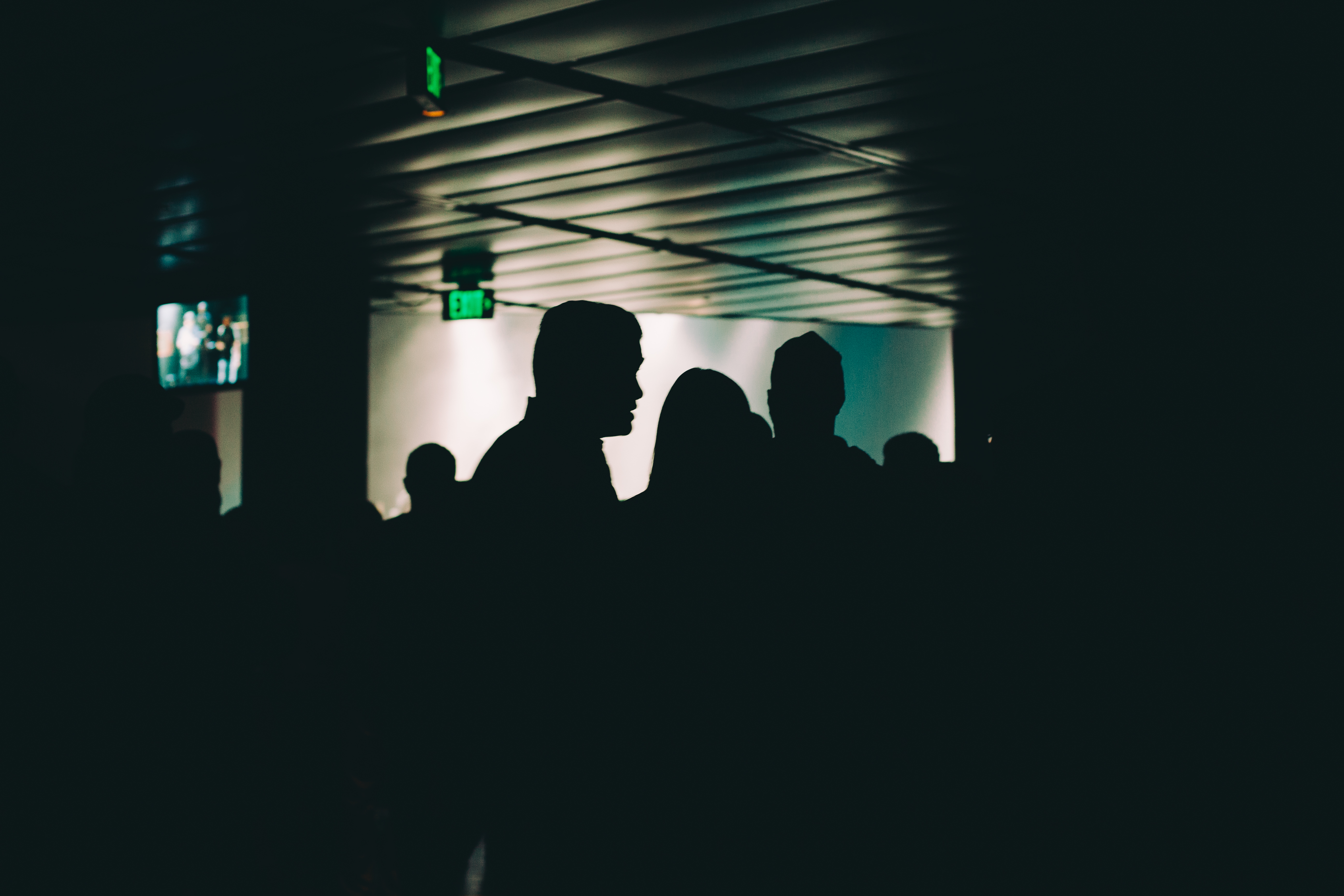 Dark silhouettes of concert attendees facing toward the lit stage in an indoor music venue.
