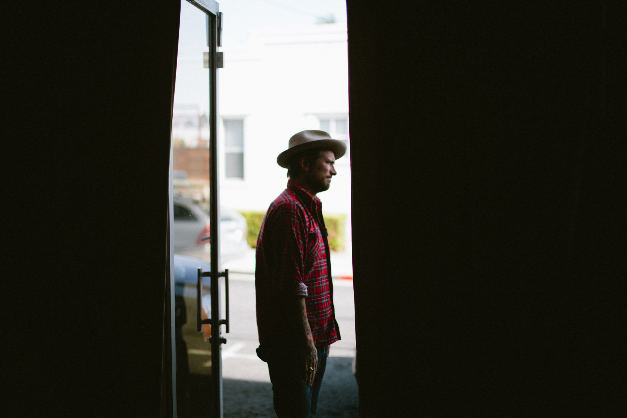 Profile portrait of musician Butch Walker wearing a fedora hat and red plaid flannel shirt standing in an open doorway looking toward the sunlight, shot with a Canon 5D Mark II.
