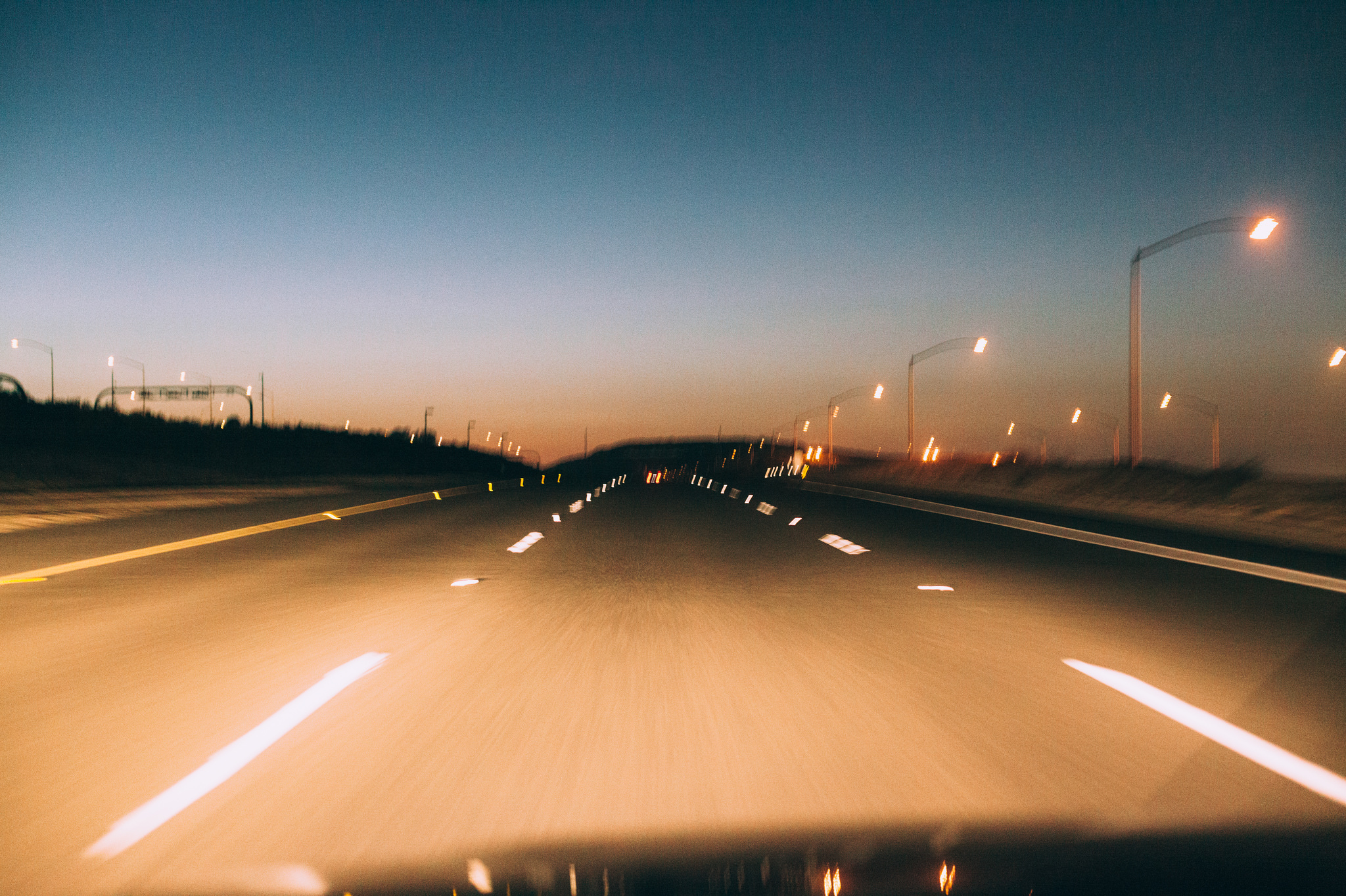 Motion blur view looking through a car windshield driving along an empty highway at dusk lined with glowing streetlights under an evening sky, shot with a Canon 5D Mark II.
