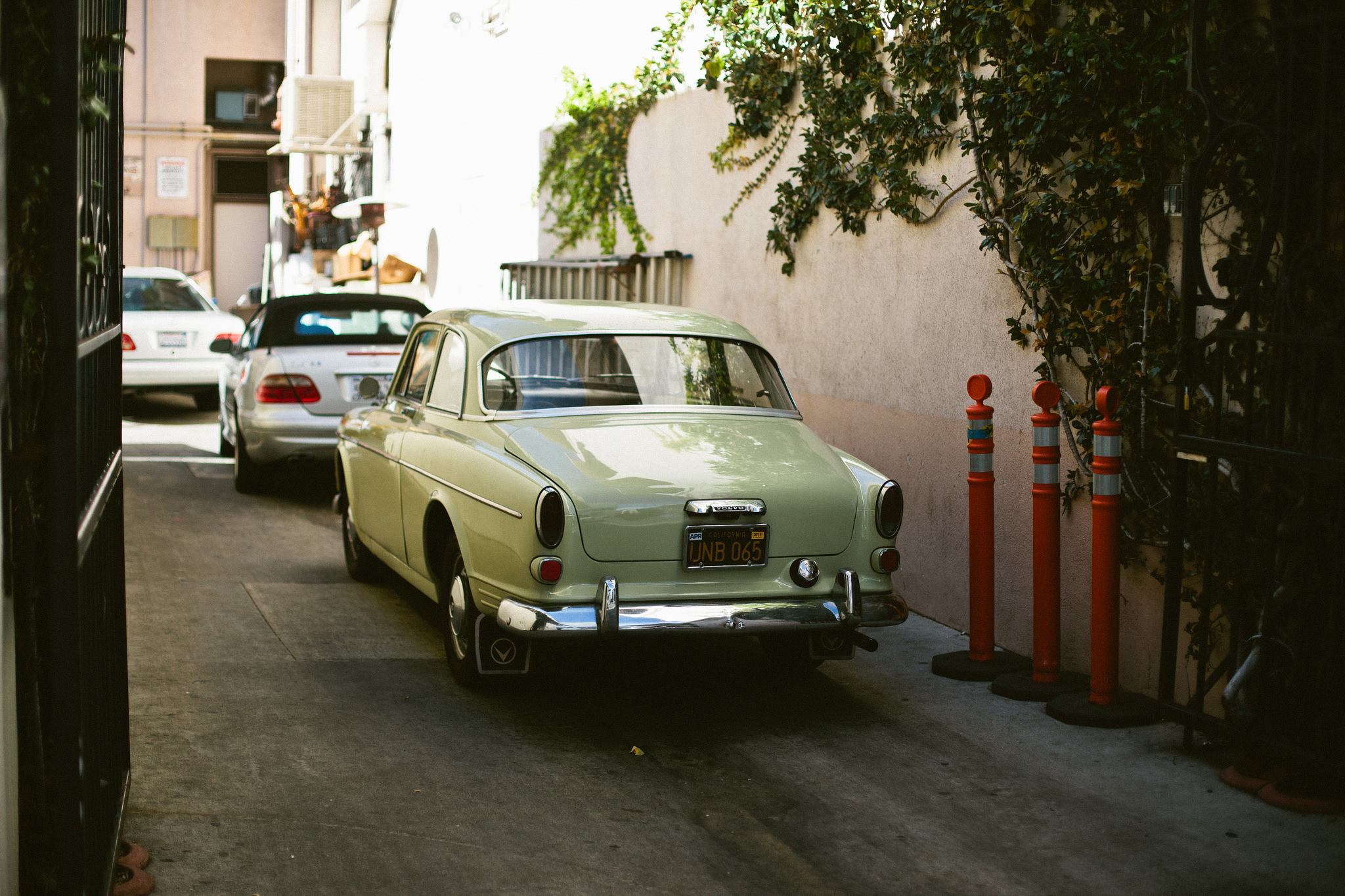 Rear three-quarters view of a vintage mint pale green Volvo Amazon coupe parked in an alleyway beside an ivy-covered wall and orange bollards, shot with a Canon 5D Mark II.