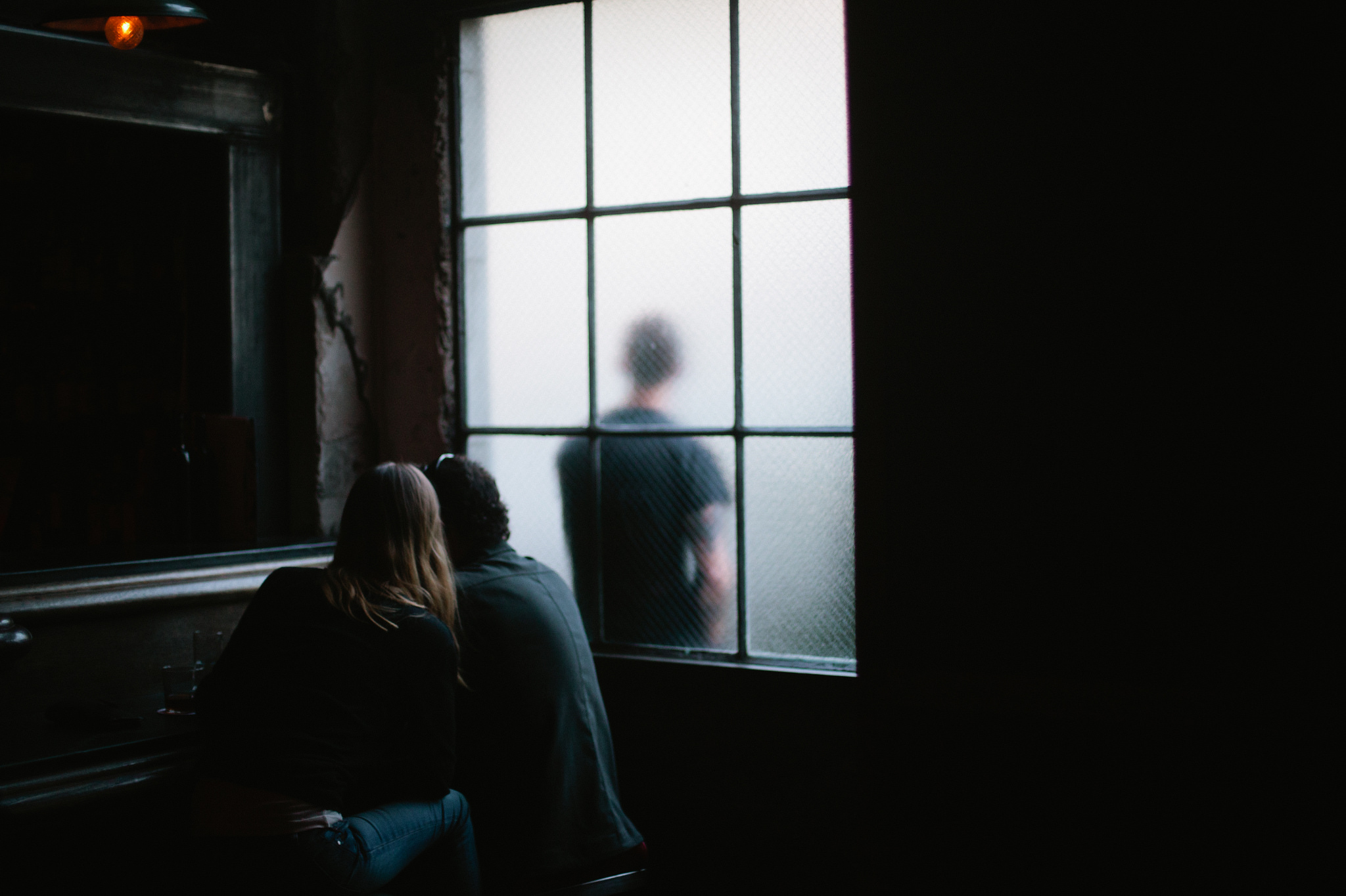 Couple silhouetted by a large frosted window in a moody bar, looking toward a figure standing outside.
