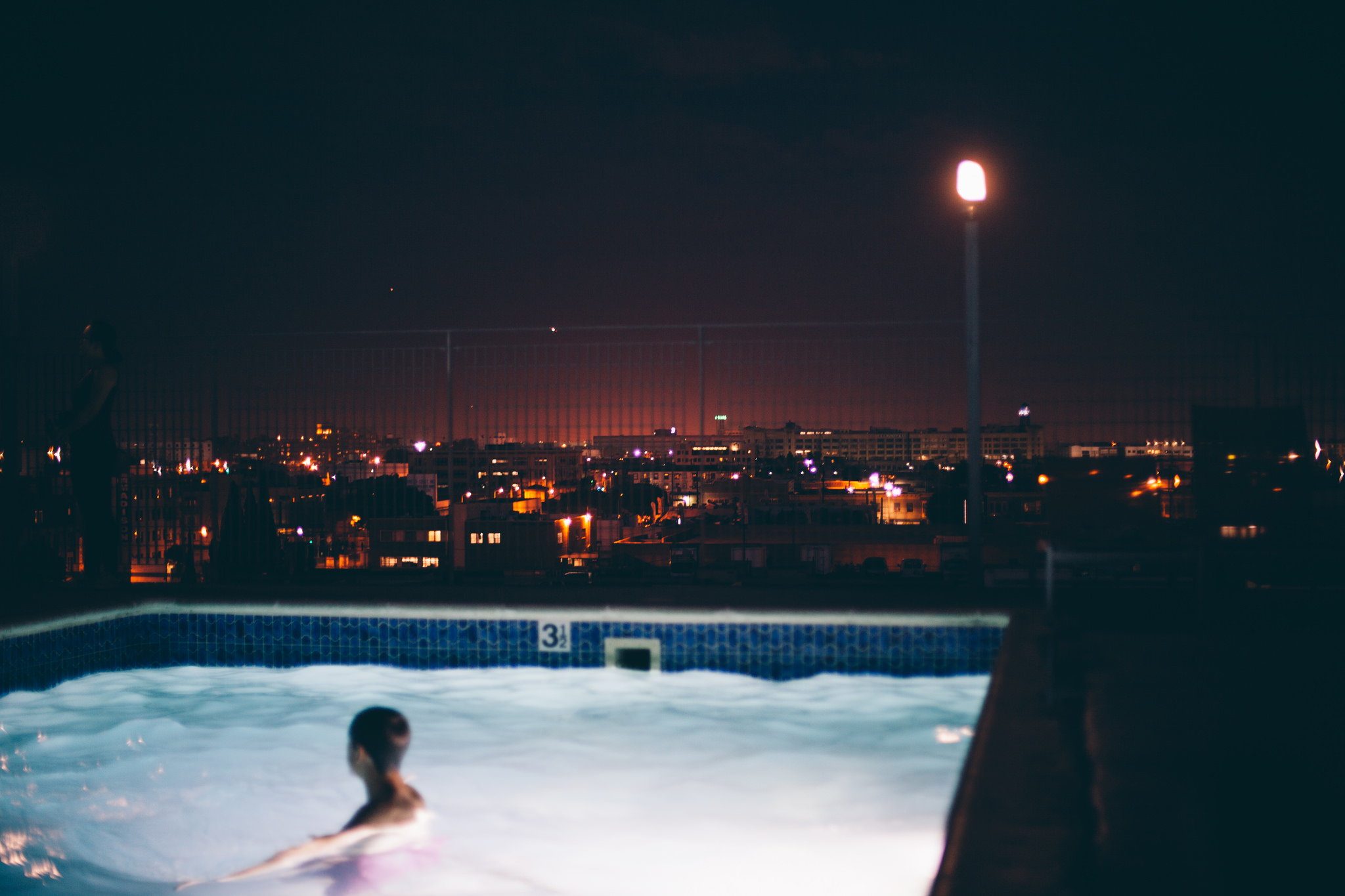 Swimmer in a glowing rooftop pool overlooking a sprawling city skyline illuminated by night lights.