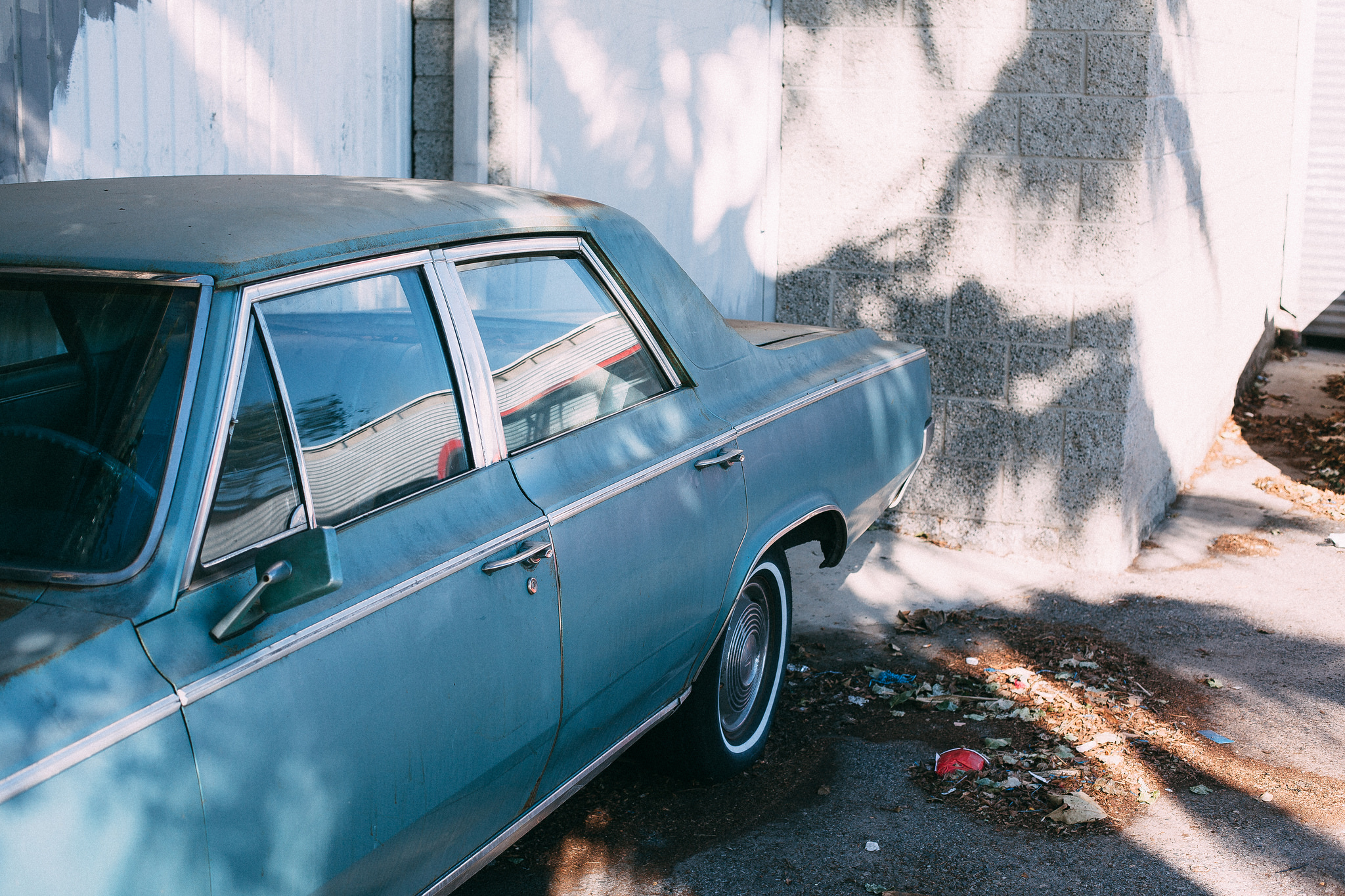 High-angle view of the rear quarter and side profile of a vintage dusty turquoise blue sedan parked in an alley beside a textured block wall with tree shadows, shot with a Canon 5D Mark II.