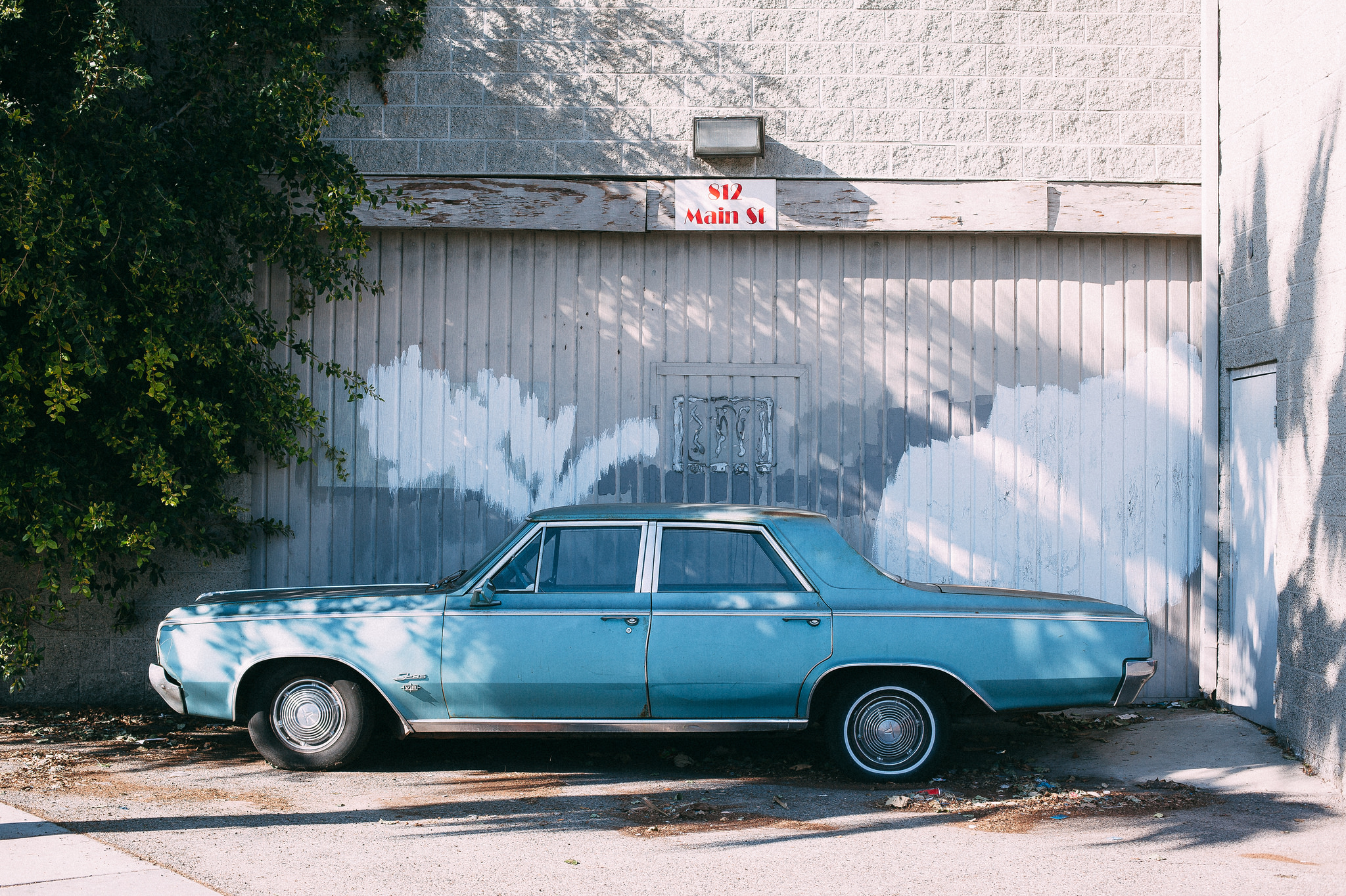Full side profile of a vintage dusty turquoise blue sedan parked outside a building with white painted garage door and 812 Main St sign, shot with a Canon 5D Mark II.