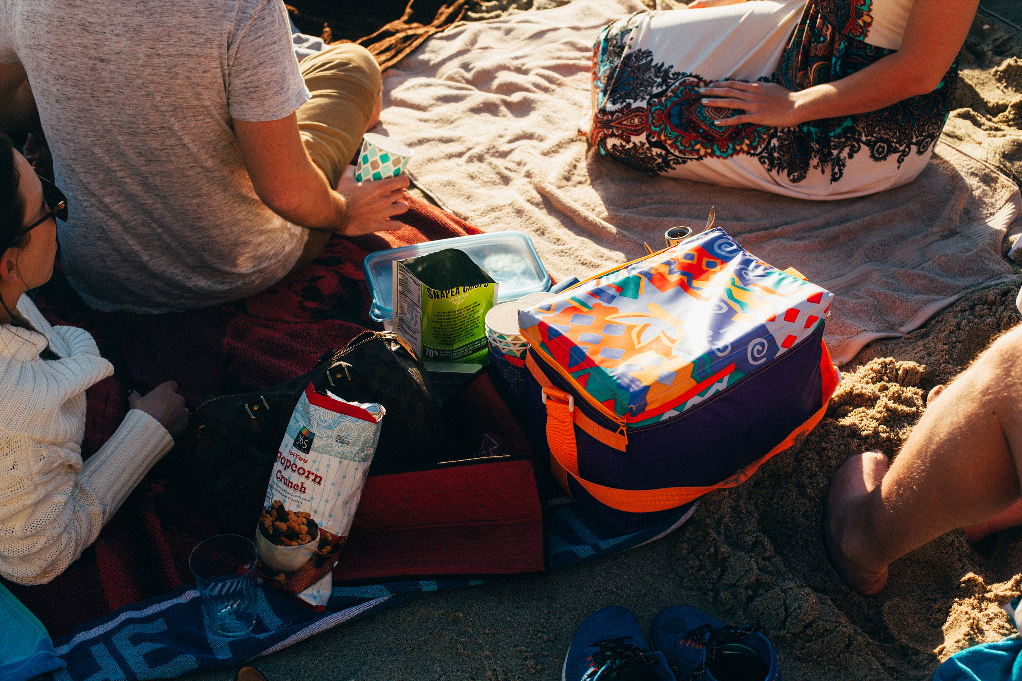 Friends having a beach picnic on blankets with snacks, drinks, and a colorful cooler bag on the sand.