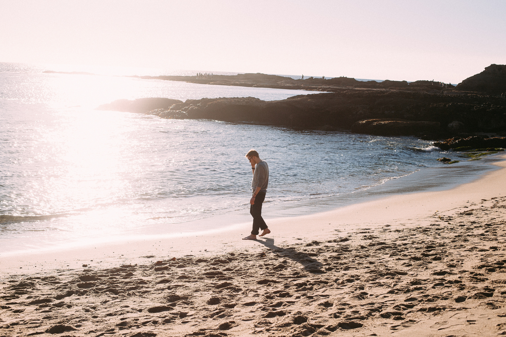 Man walking barefoot along foaming ocean surf on a sandy California beach under hazy sunlight.